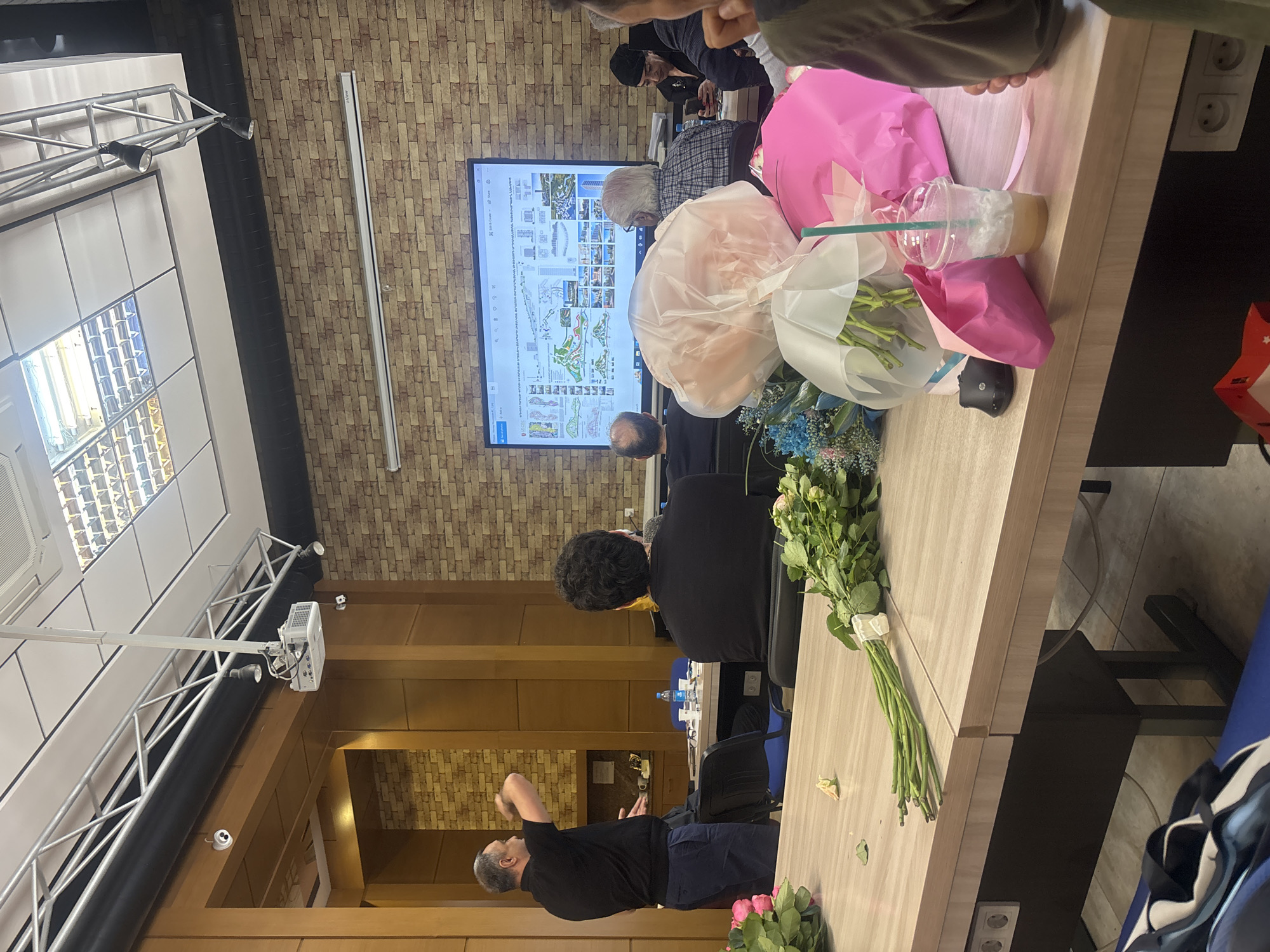 The image captures an indoor scene in Yerevan, Armenia, showing a meeting or presentation in progress. A group of people are seated at a long conference table covered with bouquets of flowers in pink and white wrapping, a drink, and other personal items. A large screen at the end of the table displays architectural designs or urban planning documents with textual information. A man stands near the table, gesturing as if explaining something. The room has brick and wood paneling, and a lighting rig is visible near the ceiling. The general atmosphere suggests a professional gathering.