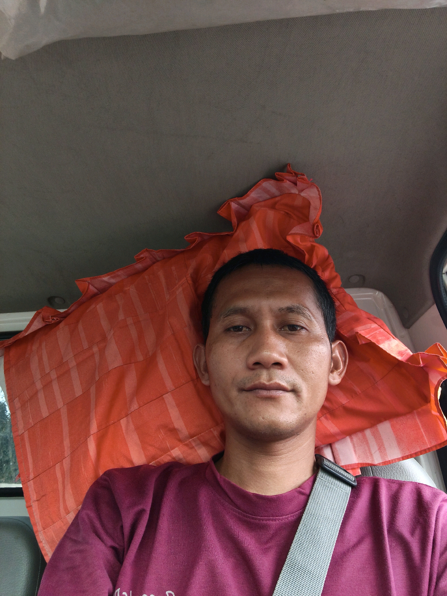 The image shows a selfie of a man taken from inside a vehicle, likely a truck cab, in Bandar Lampung, Indonesia. The man is facing the camera and appears to be wearing a maroon t-shirt. A grey seatbelt is visible across his chest. Behind him, a bright orange piece of fabric with white stripes is draped, possibly serving as a makeshift curtain or cover. The interior of the vehicle, including the ceiling and part of a seat, is visible. The overall lighting suggests it is daytime.