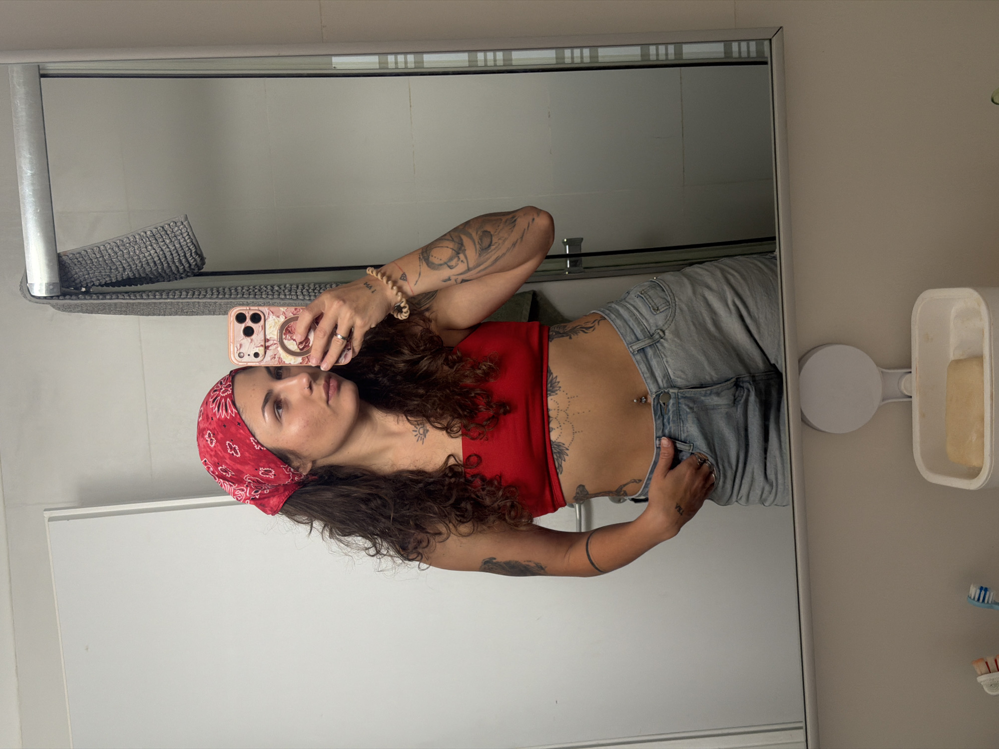 A woman stands in a bathroom, reflected in a mirror. She is wearing a red bandana, a red top, and jeans. She holds a phone, presumably taking a selfie. The bathroom features a mirror, a towel rack with a gray towel, a soap dish with a bar of soap, and toothbrushes. The location is inferred based on the implied region, but is not directly visible in the image.