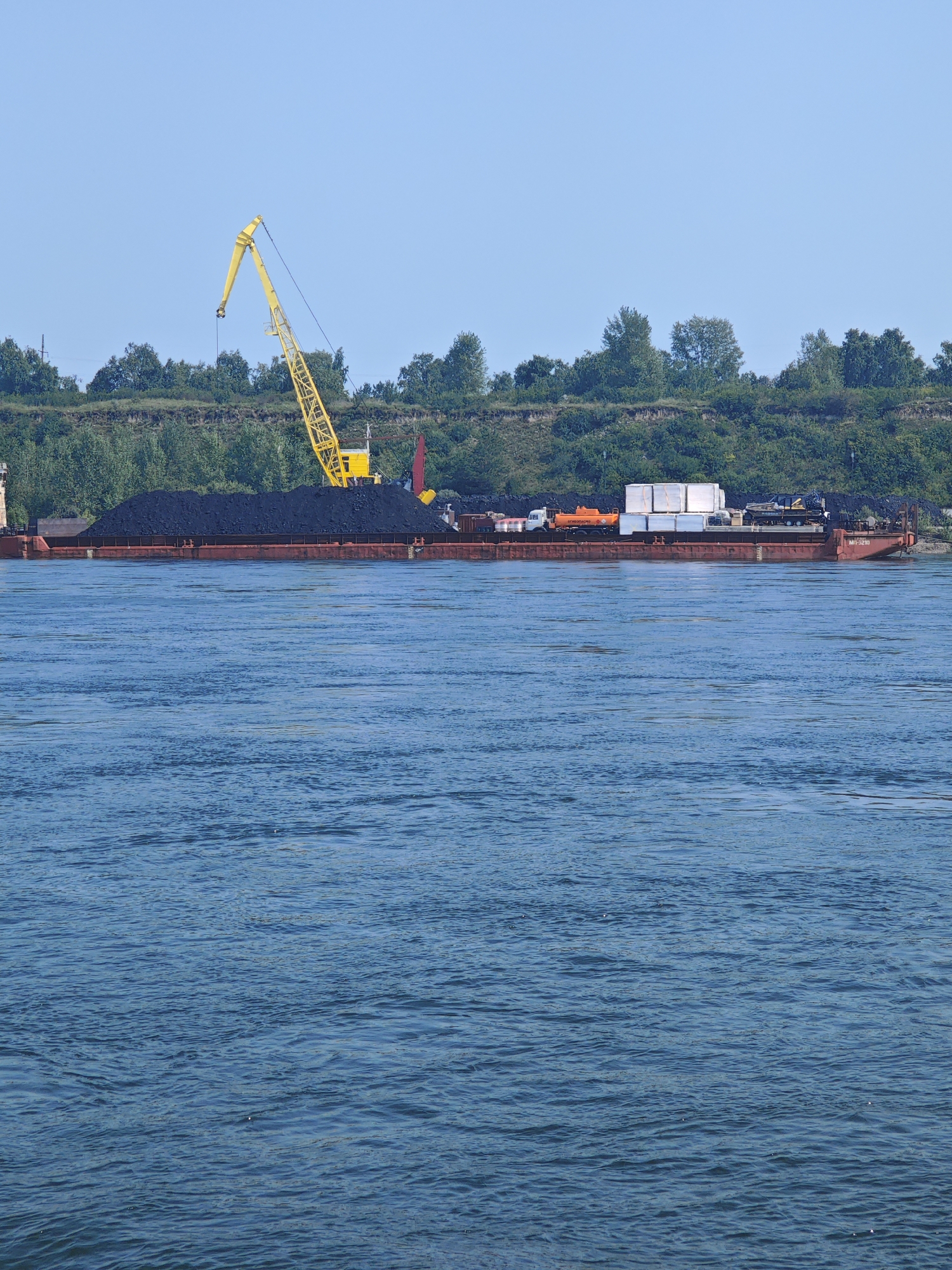 A wide, choppy, blue river occupies the foreground of the image. In the middle ground, a barge is moored near the shore. The barge is laden with a large pile of dark material, likely coal. A yellow crane is positioned on the shore behind the barge, with its arm extended over the cargo. Beyond the crane, a tree-lined embankment rises, forming the background of the scene. The sky is a clear, pale blue. The location is Berezovka, Russia.