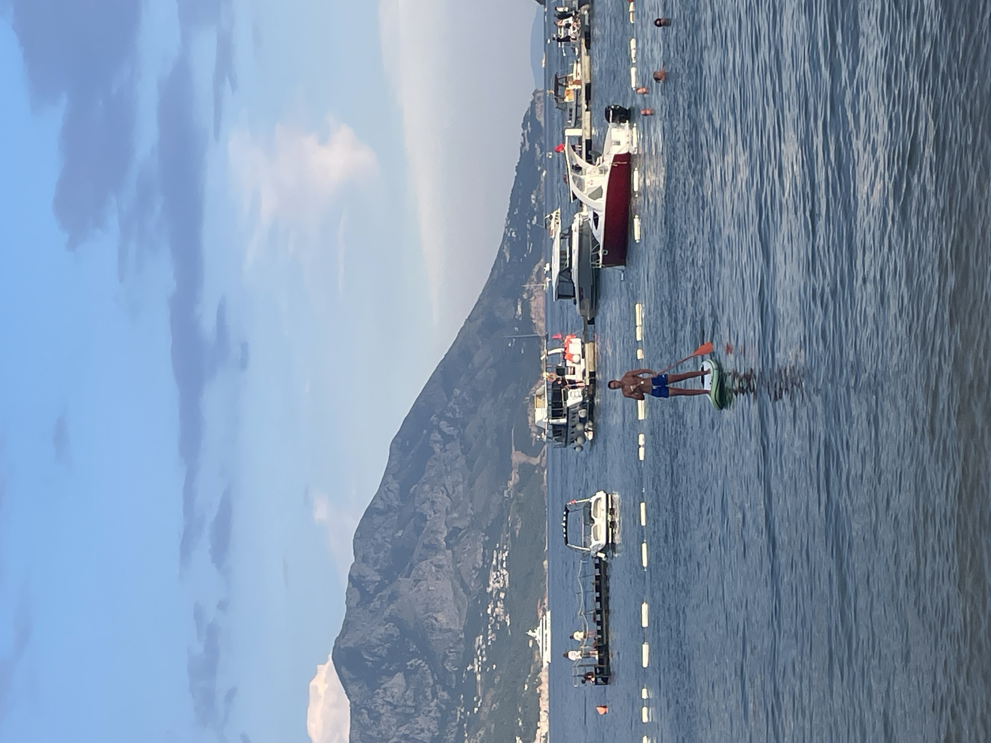 The image captures a serene coastal scene in Budva, Montenegro, likely during the late afternoon or early evening given the soft light. The dominant feature is the tranquil, rippling blue water of the Adriatic Sea, stretching towards a majestic, tree-covered mountain that rises in the background, dotted with white buildings of a coastal town.
In the foreground, a man is engaged in paddleboarding. He is standing on a light green paddleboard and holding an orange paddle, his body positioned in a relaxed stance as he glides across the water. He is wearing blue swim shorts.
Several boats are anchored or docked along the coastline, adding to the maritime atmosphere. A striking red and white boat is prominently featured, along with other smaller vessels. A long, dark pier with white lights extends into the water, and there are several buoys marking a path or boundary in the sea. The sky above is a soft blue, with scattered, wispy clouds that add texture to the overhead expanse.
The overall mood is peaceful and idyllic, evoking a sense of leisure and relaxation in a beautiful Mediterranean setting. There is no visible text in the image.