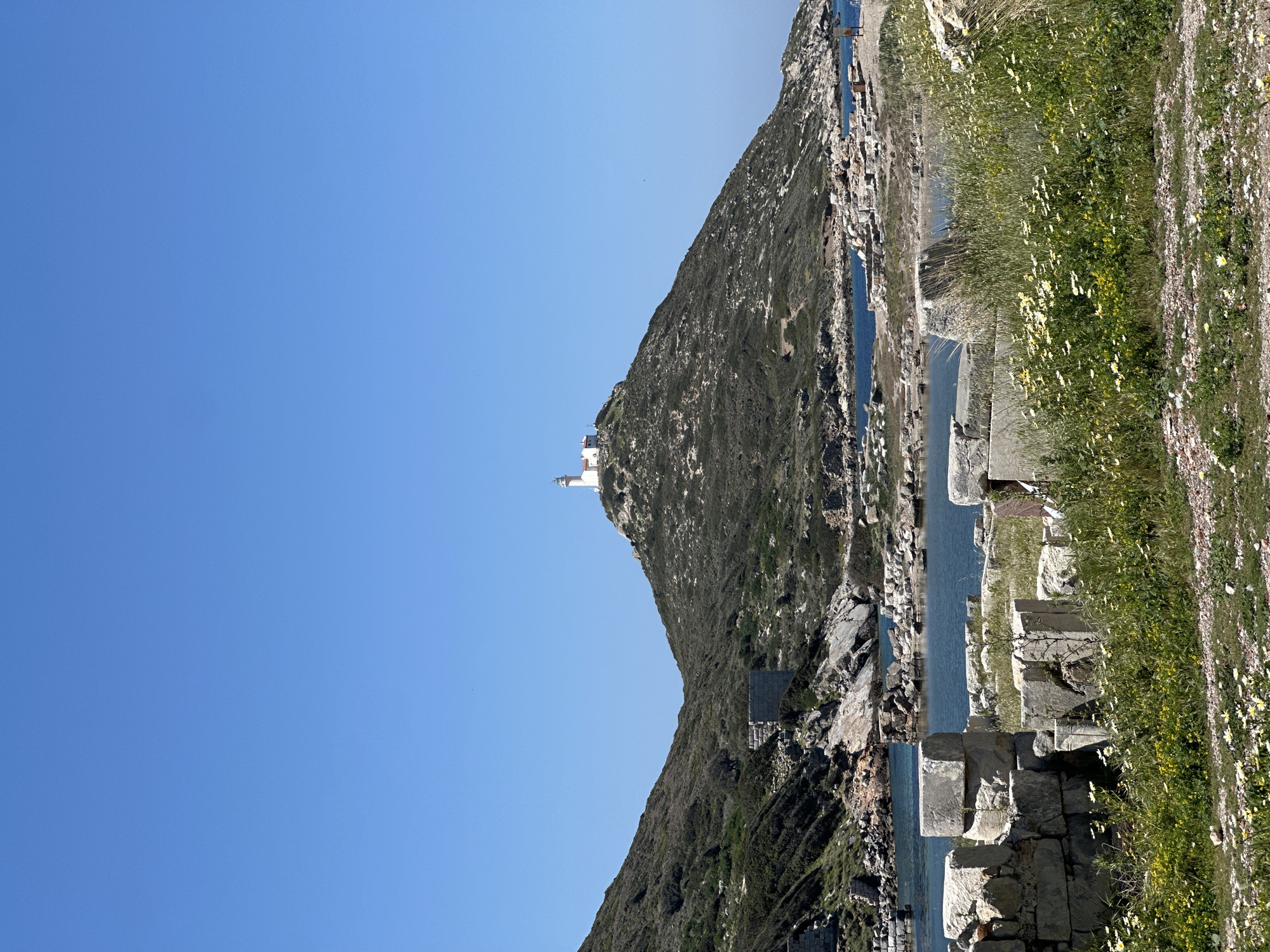 A landscape photograph depicts a hill under a clear, blue sky. A white lighthouse stands at the summit of the hill. The hill's slope descends sharply towards a narrow channel of water. Along the water's edge, there are partially submerged stone structures and walls. The adjacent shoreline is covered in greenery and wildflowers. This is likely located near the ancient city of Knidos in Datça, Turkey.