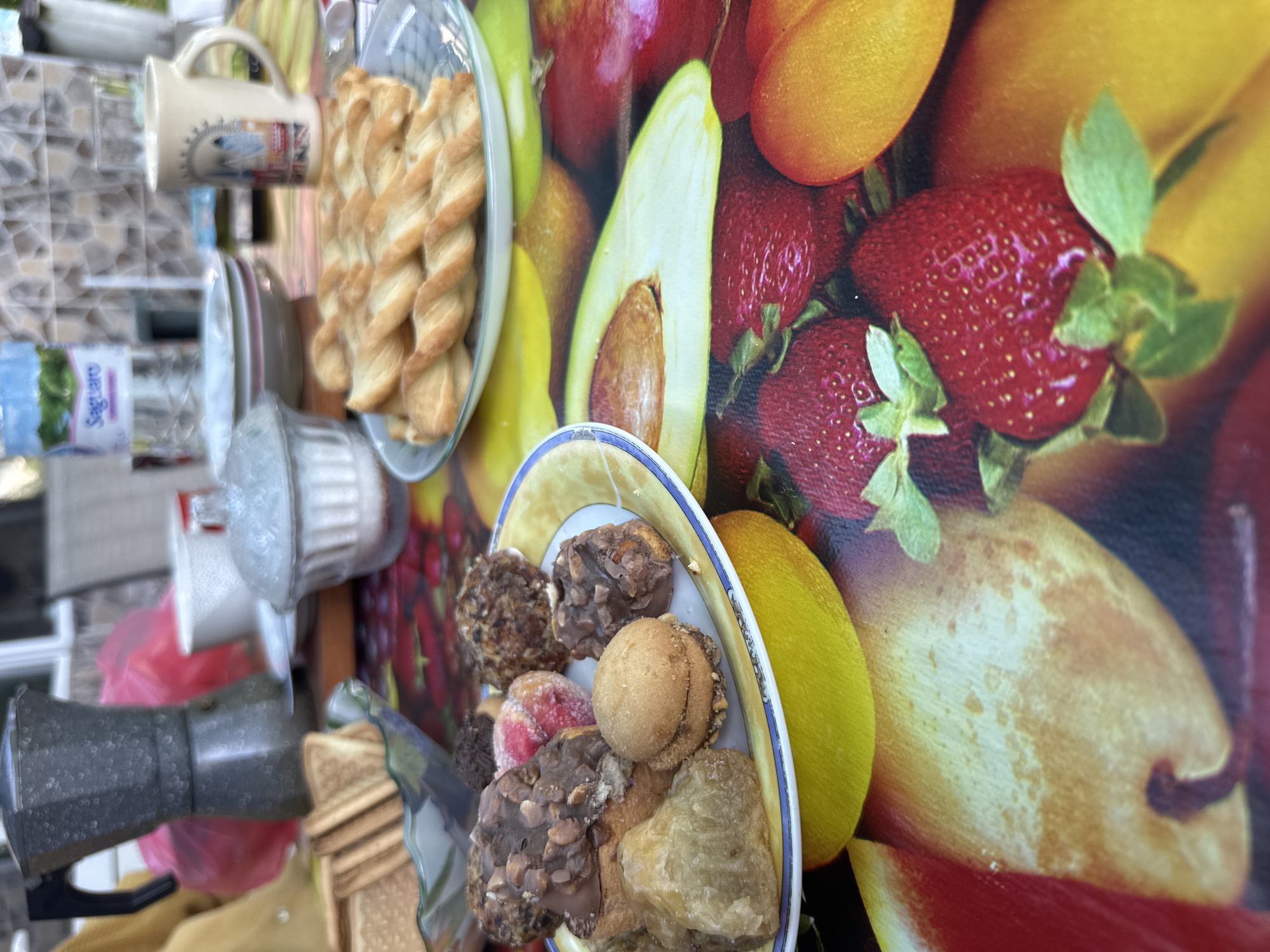 The image shows a breakfast scene with various items arranged on a table with a fruit-themed backdrop. The table appears to be set on a surface with what looks like tile, possibly a kitchen counter. A plate of twisted bread or pastry sits atop the surface along with a cup. Another plate holds a variety of small cakes and pastries. There is a coffee maker, a container with powdered sugar, a bottle of water, and some slices of toast. The background features a decorative surface displaying images of fruits like strawberries, apricots, avocado, and watermelon. There is no evidence to confirm or deny the location in Novoselets, Bulgaria, is correct. No people are directly visible.