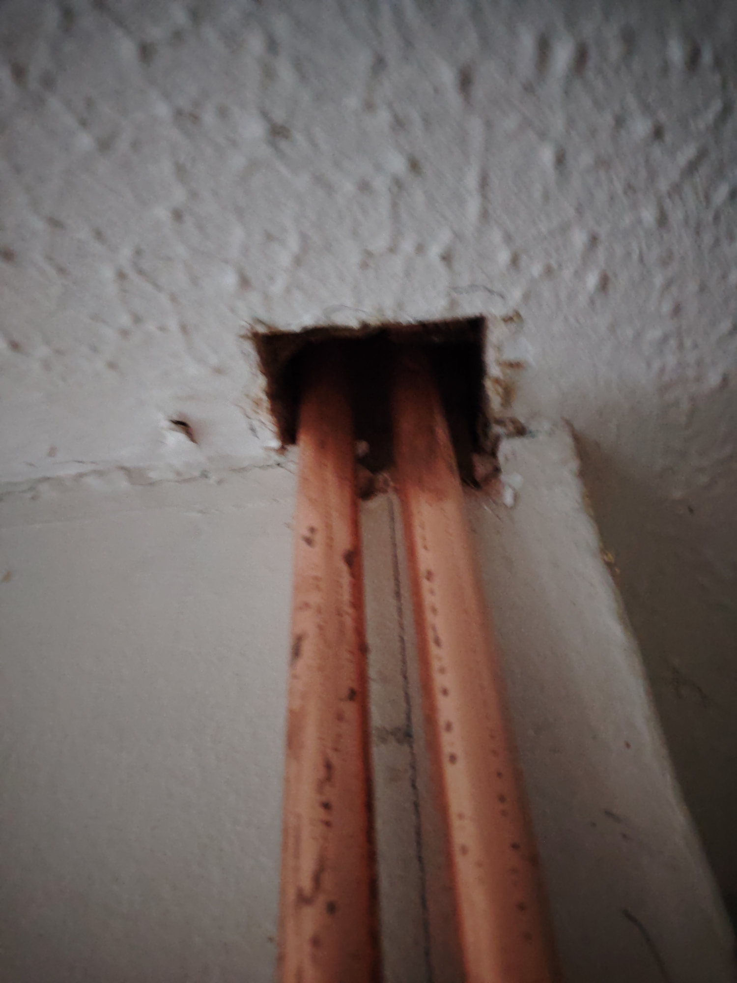 An interior, low-angle, close-up shot shows two copper pipes emerging from a roughly square opening in a textured, white ceiling. The pipes are parallel and vertical, with visible circular markings or imperfections along their length. The opening in the ceiling appears to be a hole cut into plaster or a similar material. The surrounding ceiling has a rough, popcorn-like texture. The background wall is a plain, light color, with a subtle horizontal line near the top, suggesting the edge of a different surface or a join. The lighting is somewhat dim, casting a soft shadow around the opening. The image does not contain any people, animals, or vehicles. The context of Huntingdonshire, United Kingdom, is not discernible from the visual information in the image itself.