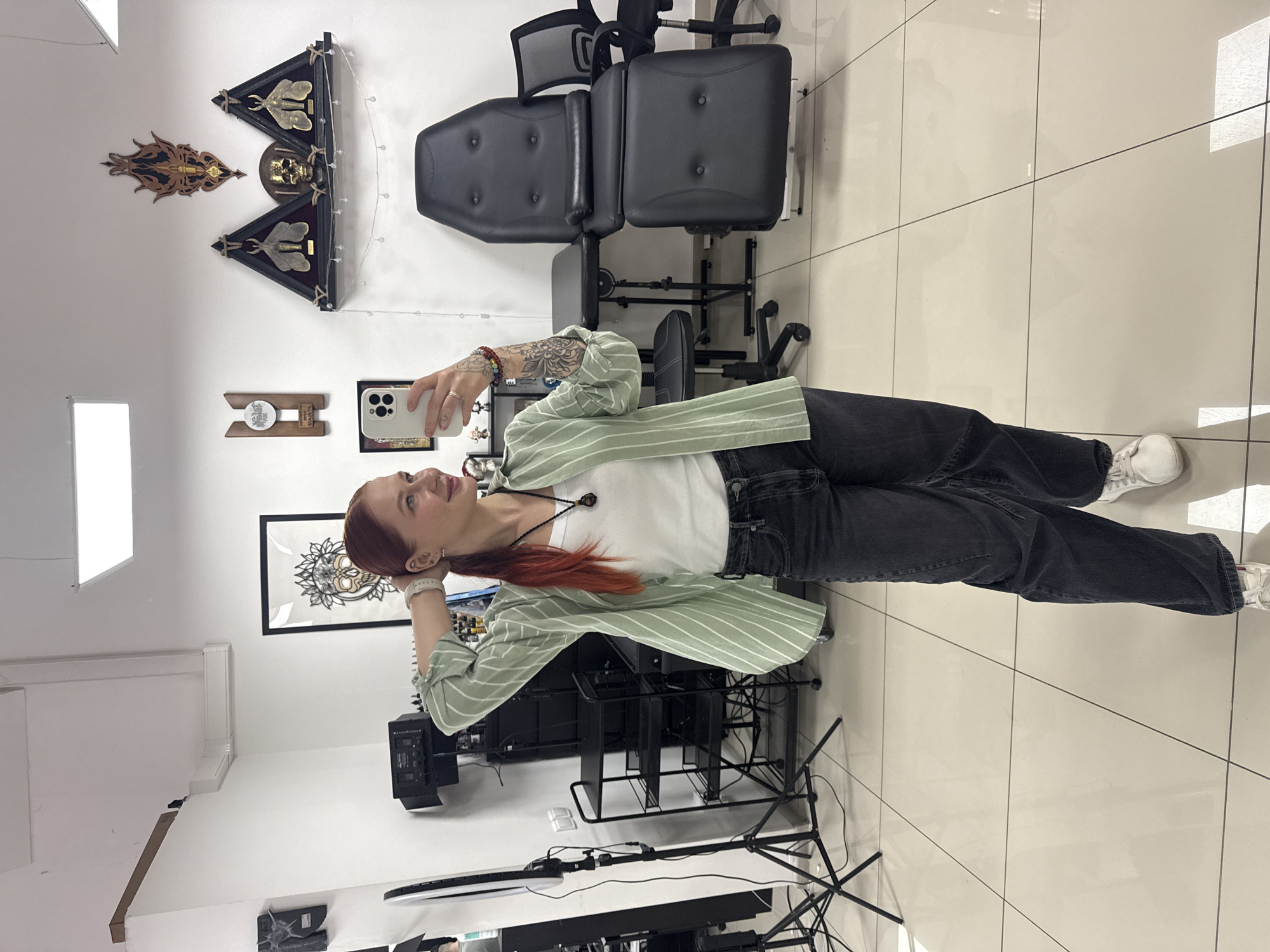 A red-haired woman is taking a selfie in what appears to be a tattoo parlor. She is wearing a white t-shirt, a light green striped jacket, and black jeans, with white sneakers. The parlor has white walls and light-colored tile floors. In the background, there is a black tattoo chair and various framed artworks and decorative items on the walls, including one with a skull. There is also computer equipment, possibly for design or business use, in the foreground.
Given the request to include the location and based on the environment suggestive of a tattoo parlor setting, it's plausible this scene is in Rostov-on-Don, Russia. However, the information isn't directly available from the photo but presumed.