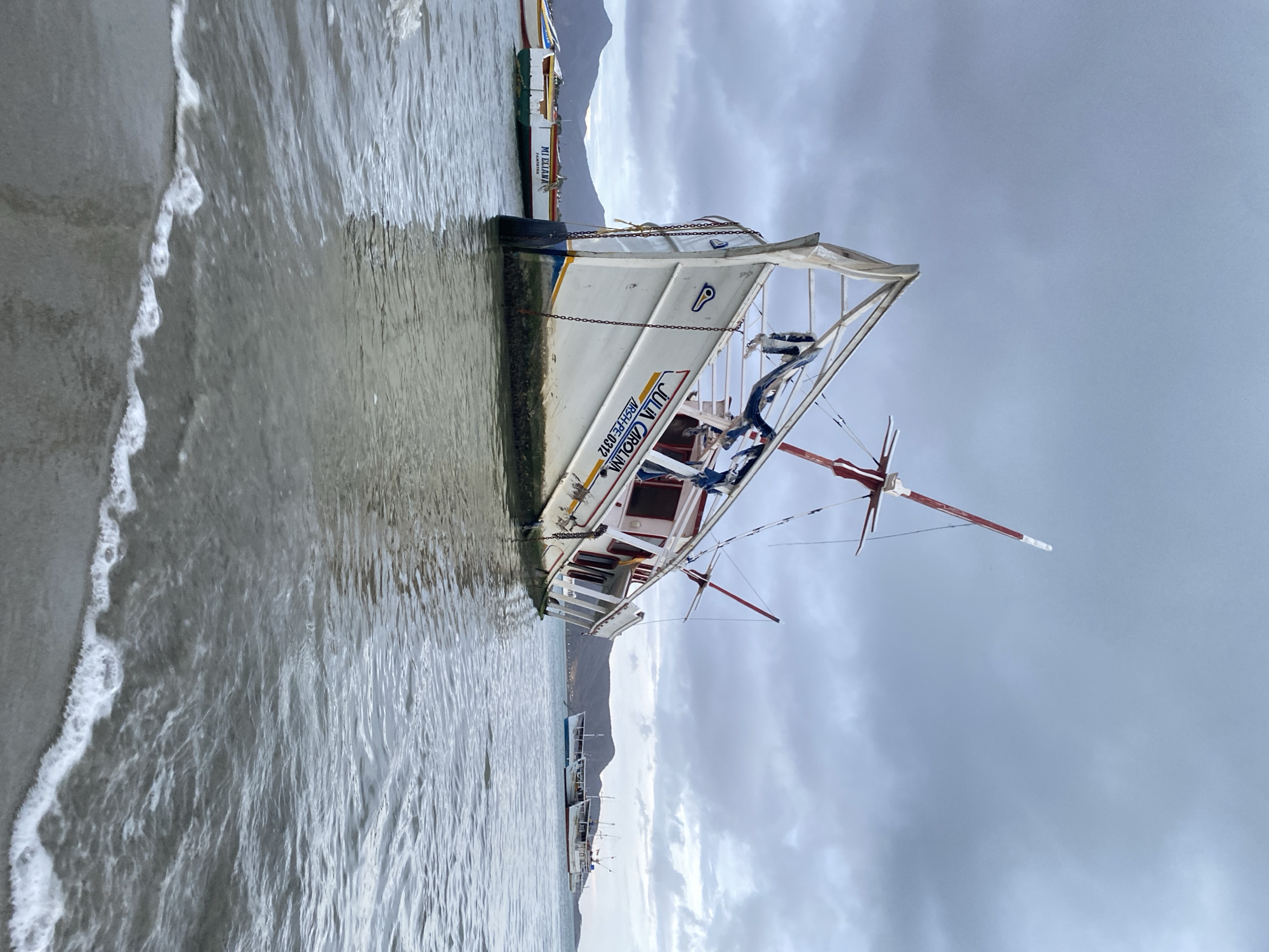 A sideways medium shot shows a white boat, named "Julia Carolina" and "Archpi.pe 0312," resting partially submerged on a sandy beach. The water level reaches the lower hull of the boat, and a wave washes up the beach. Other boats are docked in the distance. Behind the boats, there are mountains, and above, there is a cloudy sky. It's likely in Juan Griego, Venezuela, where similar scenes are common.