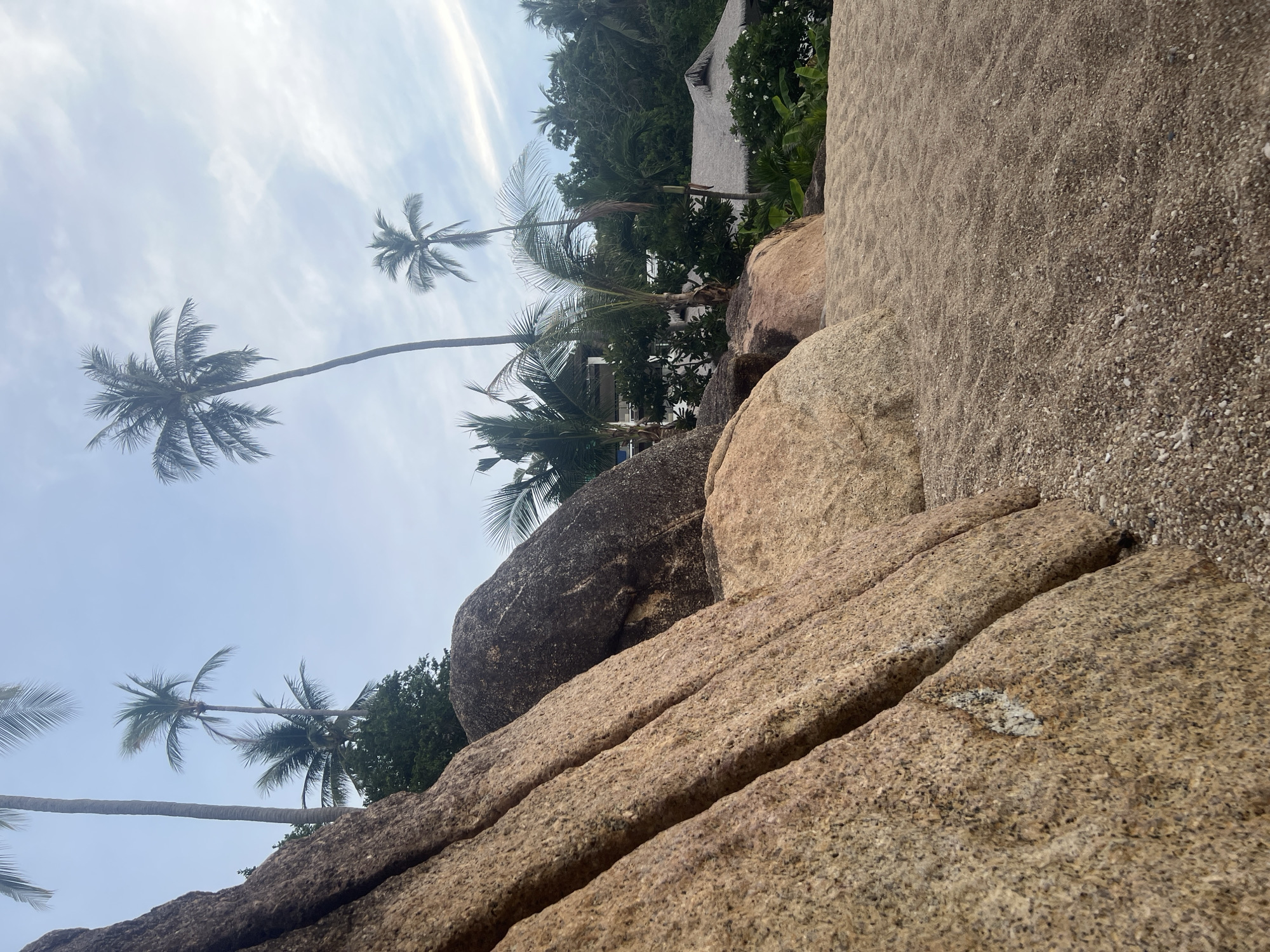 The image depicts a tropical beach scene in the Ko Samui District, Thailand. In the foreground, large, textured granite rocks dominate the left side, with their rough surfaces showing variations in color from light brown to darker gray. A sandy beach stretches to the right, with small pebbles and grains of sand forming gentle undulations. In the background, lush green palm trees and other tropical vegetation are visible. Several thatched-roof buildings, likely part of a resort or residential area, are nestled among the trees. The sky is a pale blue with wispy white clouds, suggesting it is either early morning or late afternoon. There are no people or active events depicted in the image.