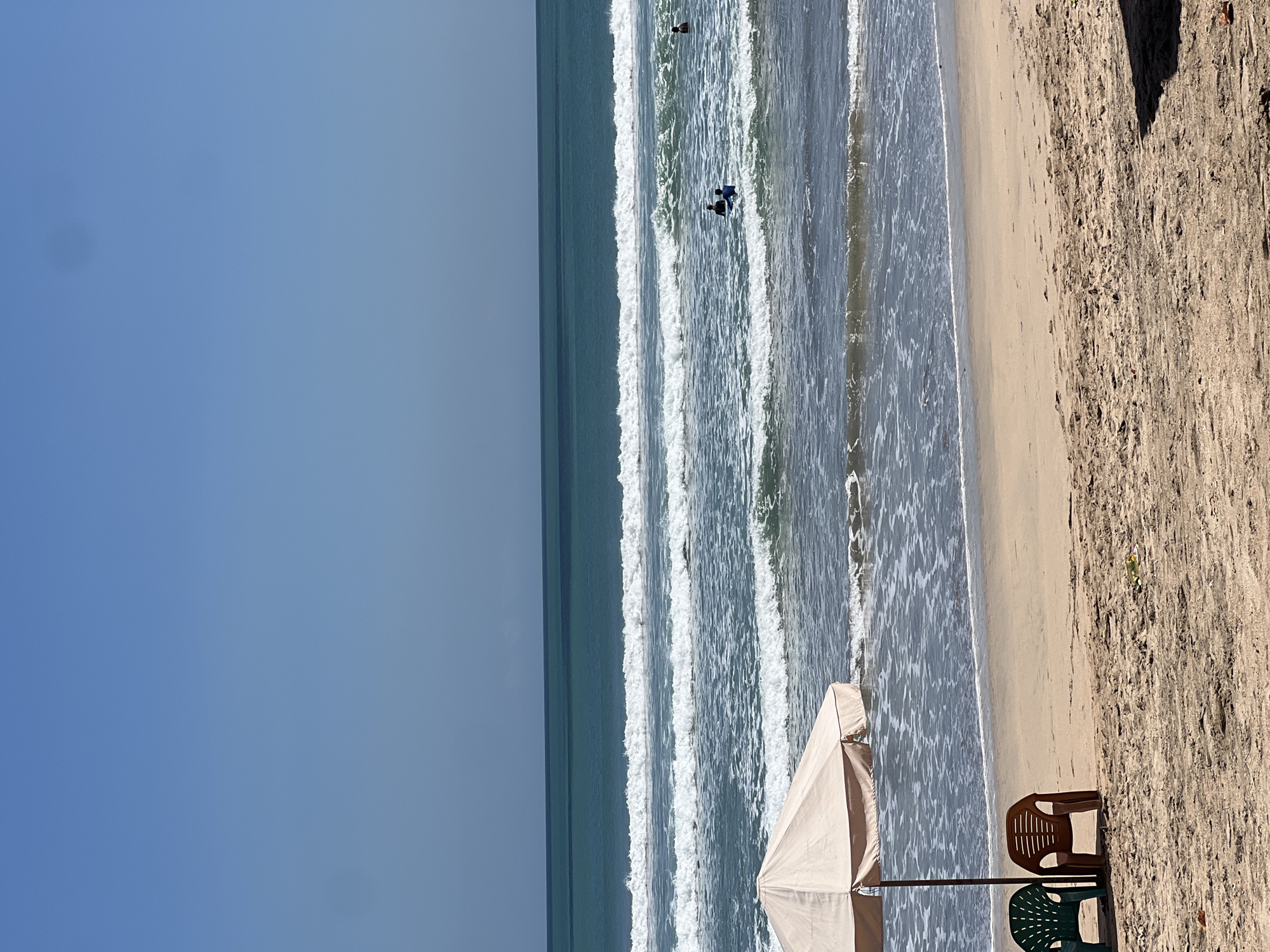This image depicts a serene beach scene in Kuta, Indonesia, under a clear blue sky. In the foreground, two plastic chairs, one green and one brown, are positioned beneath a beige beach umbrella. The sandy beach stretches towards the ocean, where gentle waves are breaking. Several surfers are visible in the water, enjoying the surf. The ocean transitions from a lighter shade of blue near the shore to a deeper teal further out, with distinct lines of breaking waves creating a sense of movement and tranquility. The overall atmosphere is one of peaceful relaxation on a sunny day.