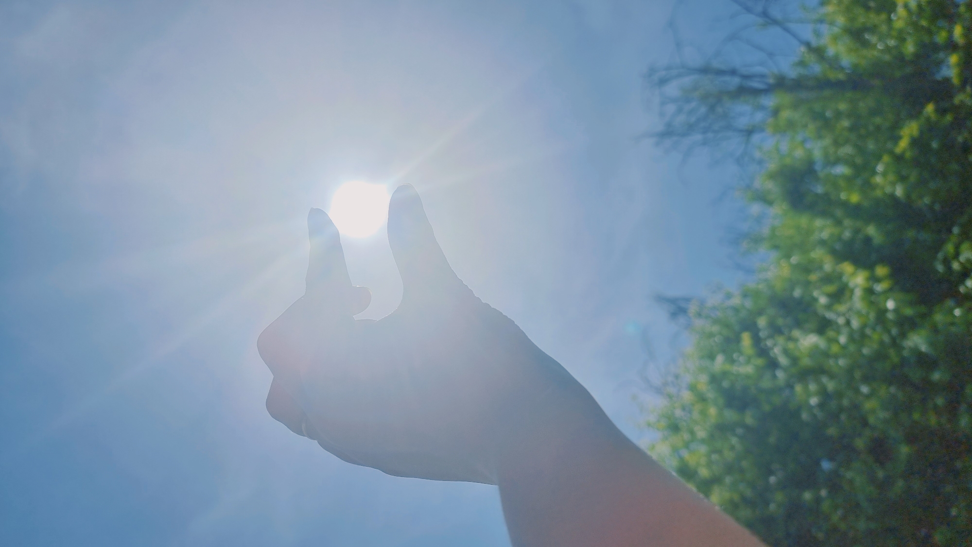 The image shows a hand obscuring the sun against a bright blue sky with some trees visible in the background. The location is presumably in Dunnae-myeon, South Korea, as requested. The sun is emitting rays of light. The hand is silhouetted and positioned between the viewer and the sun.