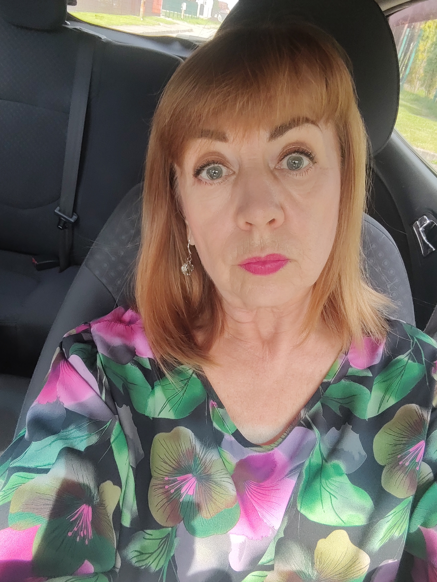 An indoor, close-up, selfie-style photograph shows a Caucasian woman with auburn hair and bangs. She is seated in the driver's seat of a vehicle. The woman is wearing a V-neck blouse with a floral pattern featuring pink, green, and yellow flowers. Her lips are painted a bright pink. She is wearing a silver earring in her visible left ear. The background includes the car's headrest, a seatbelt, and out the window, what appears to be a street scene with buildings and greenery, consistent with a residential area of Gvardeysk, Russia. The lighting suggests it is daytime.