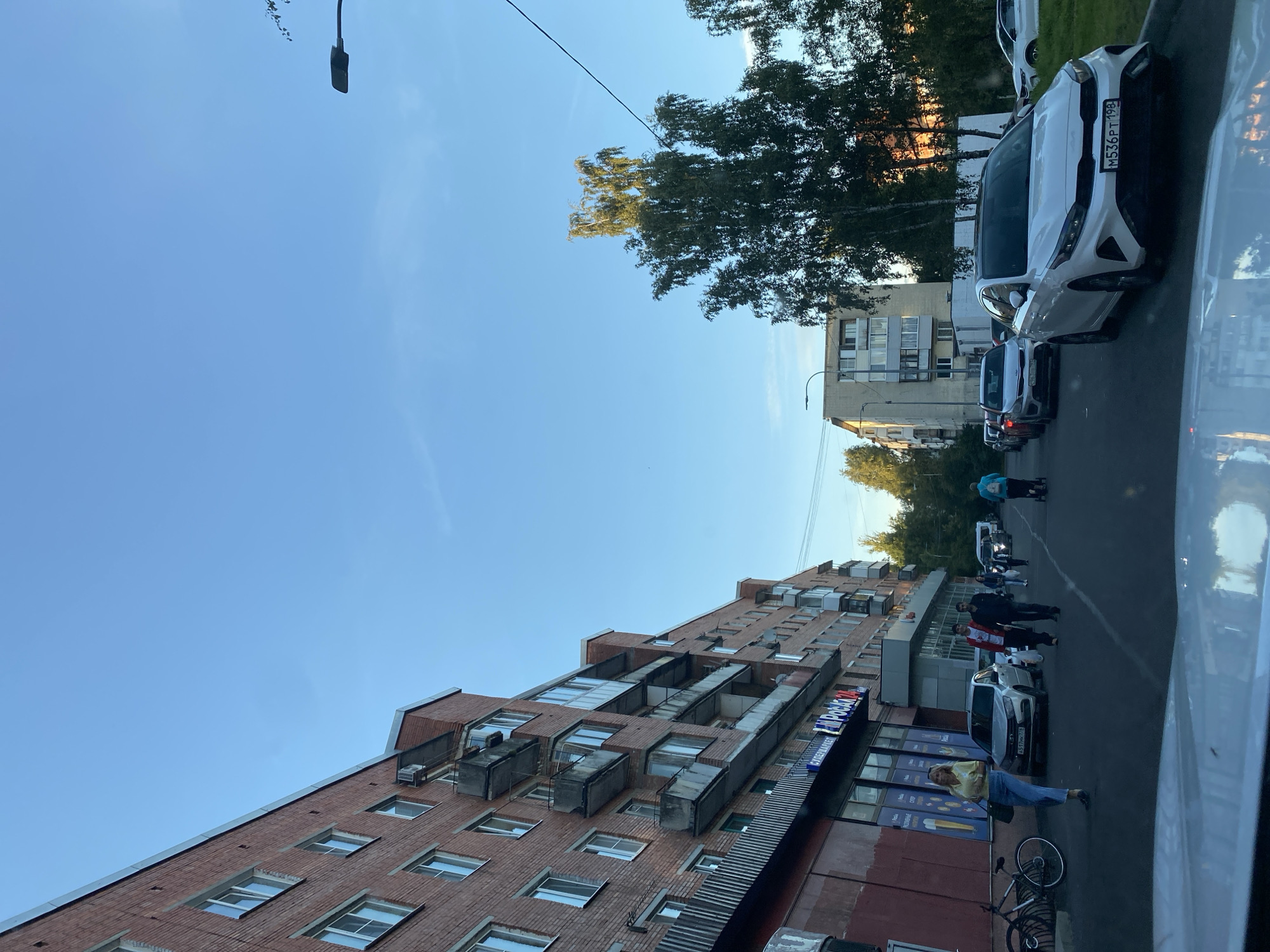The image depicts a street scene in Lomonosov, Russia, likely during daylight hours with a clear sky. A multi-story brick apartment building is visible on the left, with balconies and windows. A sidewalk runs along the building, where several individuals are walking. A bicycle is parked on the sidewalk. A white sedan is parked in the street in front of the building. Further down the street, other cars are parked and moving. Trees are present on the right side of the street, with one tall tree prominently featured. Another apartment building, lighter in color, is visible in the background. A street lamp is also visible against the sky. The perspective suggests the photo was taken from inside a vehicle.