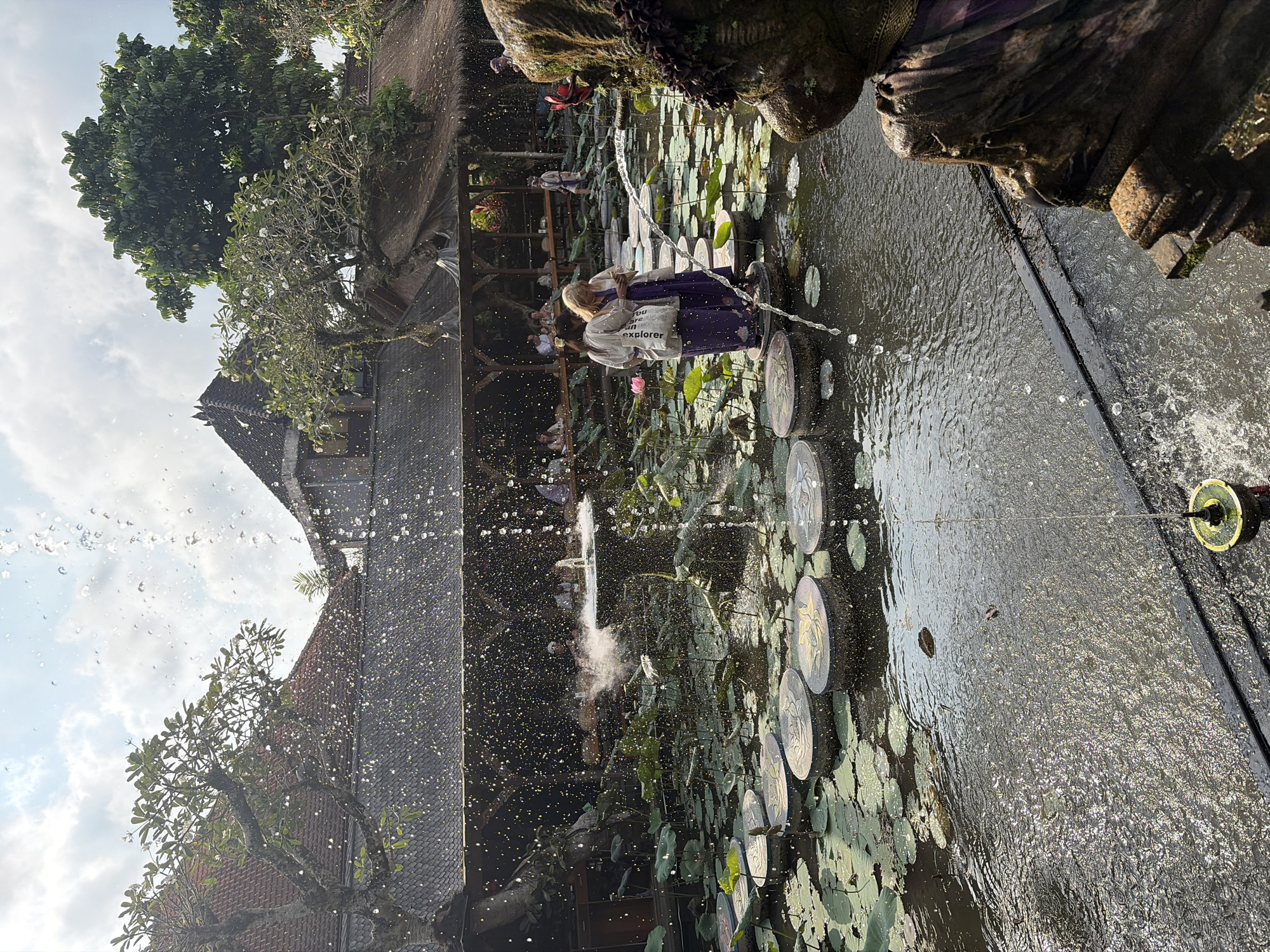 In Ubud District, Indonesia, a courtyard features a tranquil water garden. The pond, filled with lily pads, is punctuated by stepping stones, and a fountain sprays water into the air. Two women, dressed in traditional Balinese attire, stand on the stepping stones, facing away from the camera. They appear to be engaged in conversation or looking at a device. In the background, an open-air restaurant is visible, with patrons seated at tables. The architecture is characteristic of Balinese style, with thatched roofs and intricate carvings. The scene is bathed in sunlight, casting dappled shadows across the water and foliage.
