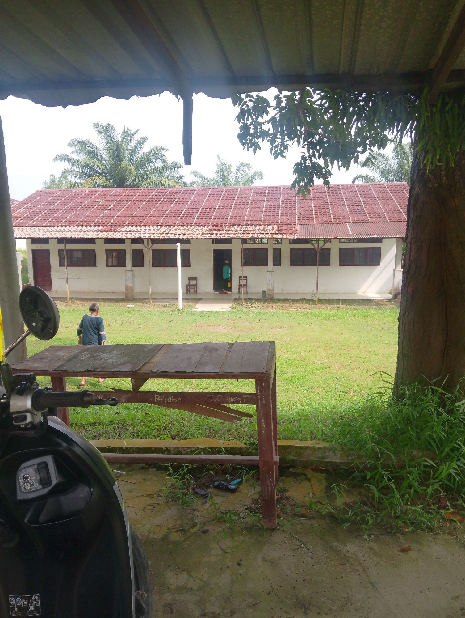 In the city of Serbelawan, Indonesia, a long, single-story building with a red tiled roof and white walls stands amidst a grassy yard. The building features multiple windows with dark brown frames and a series of identical doors. Two individuals are visible near the building entrance. One, a person wearing a blue shirt, walks away from the camera towards the building. Another individual, in a light blue shirt, stands near an open doorway.
In the foreground, a wooden table with a weathered top rests on a concrete surface. A portion of a black scooter, including its handlebars and front panel, is visible on the left side of the image. The scooter's control panel, featuring power and light buttons, is clearly depicted. The environment is lush with green grass and vegetation, including palm trees visible in the background and a large tree trunk on the right. The scene is framed by a dark, corrugated roof overhang, with natural light illuminating the area.