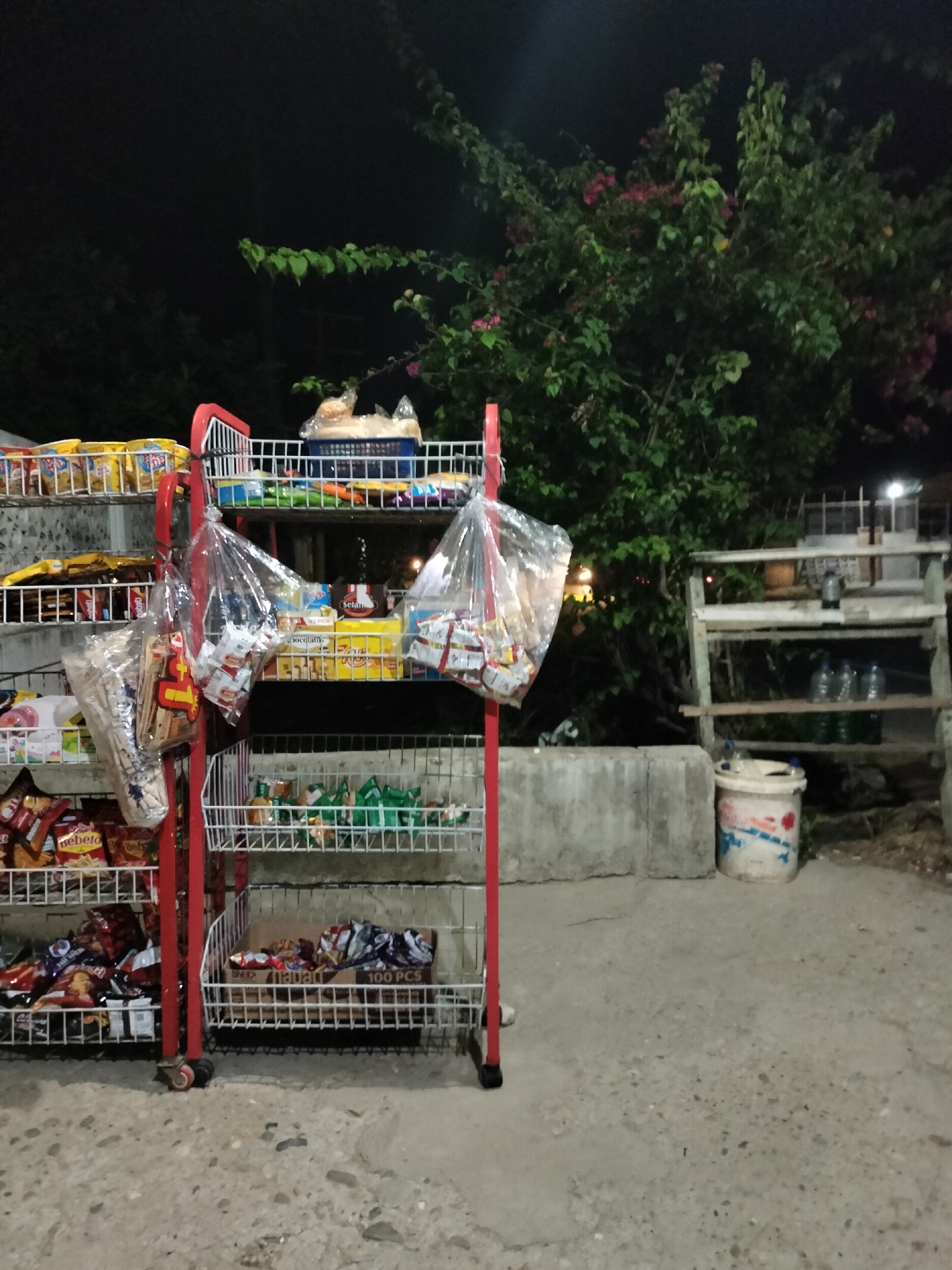 The image shows an outdoor street stall at night in Rantopanjang, Indonesia. The stall is set up with red metal shelving units filled with various packaged goods, primarily snacks and drinks. Some items are in clear plastic bags hanging from the shelves. To the right of the stall is a larger, weathered wooden shelving unit with bottles on it, and a concrete barrier. Further in the background, a lush green tree with some pink flowers is visible, along with other structures that are dimly lit. The ground is paved with concrete and gravel. There are no people visible in the image.