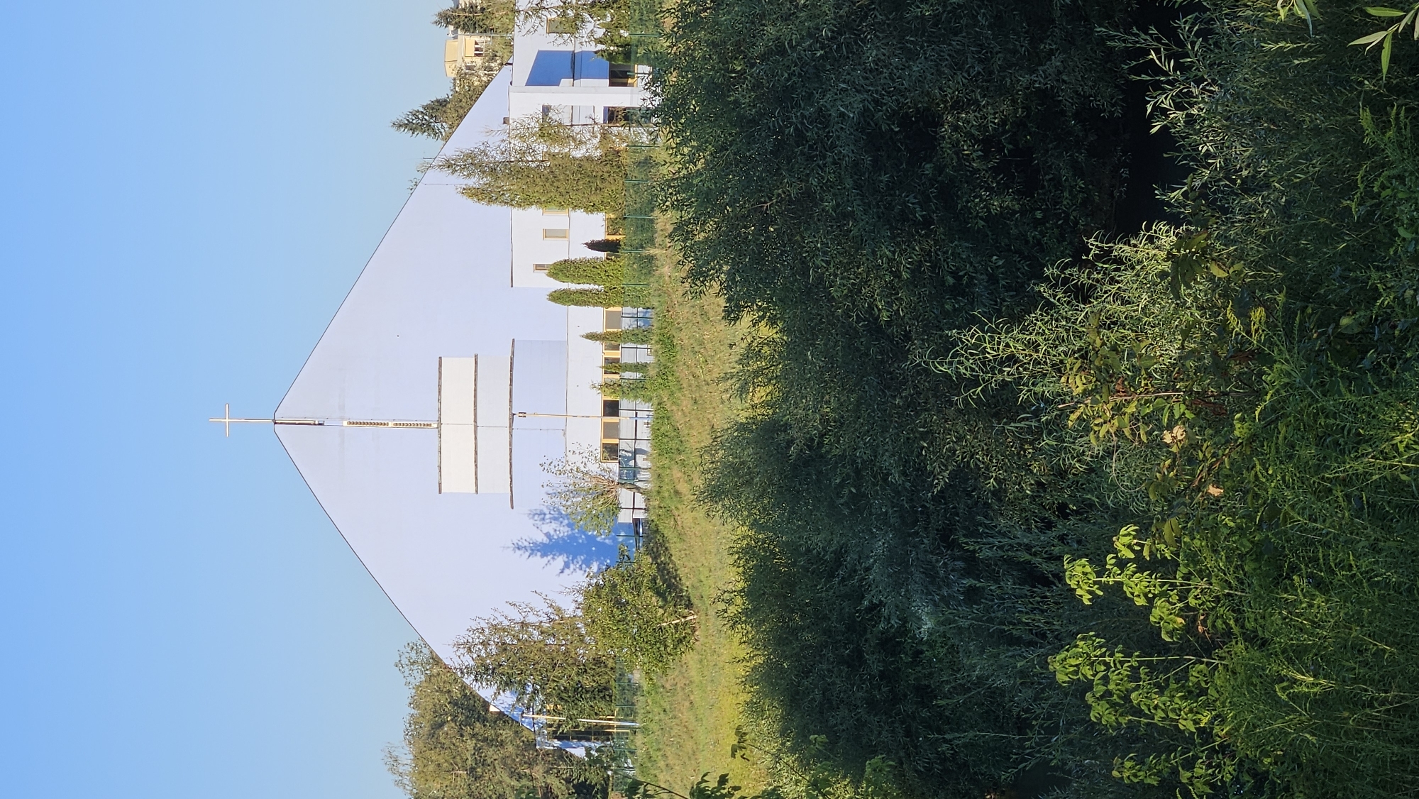 The image displays a modern, light blue church building with a prominent cross on its roof. The church is situated on a grassy hill, with a dense green bush in the foreground. The sky is clear and blue. In the background, several trees and other buildings are visible, suggesting an urban or suburban setting. Based on available information, this church is likely the Church of the Holy Spirit, located in the Sekčov district of Prešov, Slovakia. The building appears to be in good condition, and there are no people or animals visible in the frame. The lighting indicates it is either early morning or late afternoon, with the sun casting shadows.