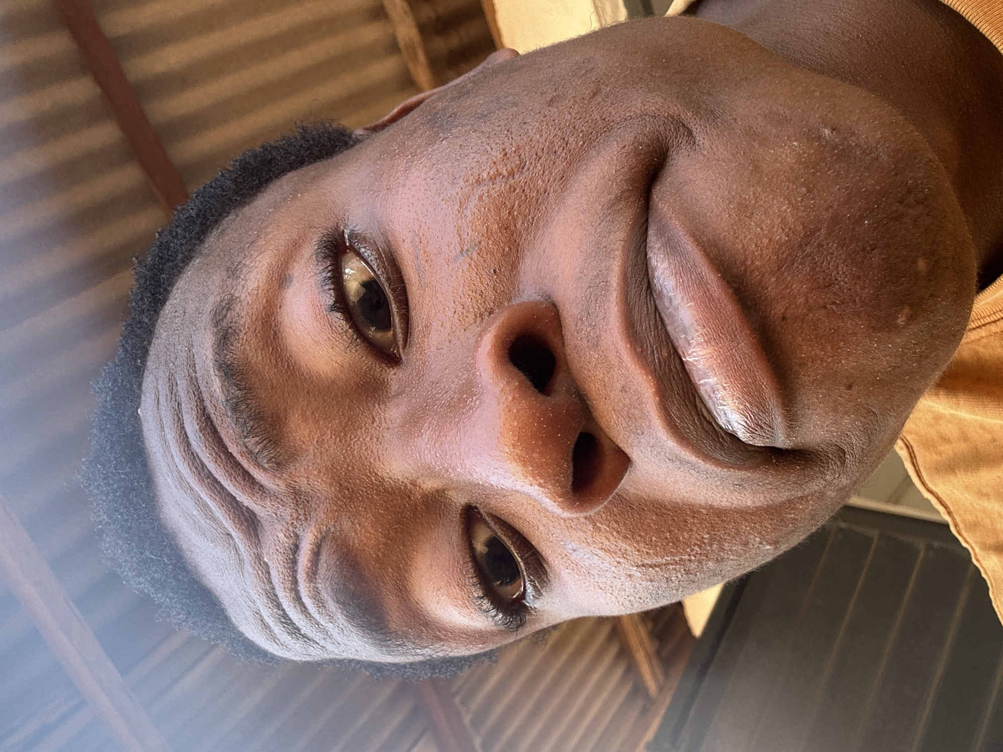 This close-up, low-angle portrait captures the face of a dark-skinned individual, likely male, with their head tilted and eyes looking directly downwards towards the camera. The image is rotated, presenting the face diagonally across the frame. The person has short, dark, tightly curled hair visible around the temples and forehead.
Their facial features include prominent eyebrows, dark brown eyes with visible eyelashes, and a well-defined nose. The lips are full, and the right corner of the mouth is subtly upturned, suggesting a slight smirk or relaxed smile. The skin on the face exhibits natural texture, including some visible pores, minor blemishes, and creases on the forehead, likely due to expression or the angle of the shot. The skin appears slightly dewy or oily, reflecting the bright light.
A hint of a light brown or tan garment, possibly a collar, is visible on the right side of the frame. The background is blurred but shows distinct corrugated metal panels, likely a ceiling or roof structure, suggesting an indoor or semi-enclosed setting such as a shed or garage. The bright, natural lighting indicates daytime, illuminating the subject's face and creating highlights in their eyes. No visible text is present.