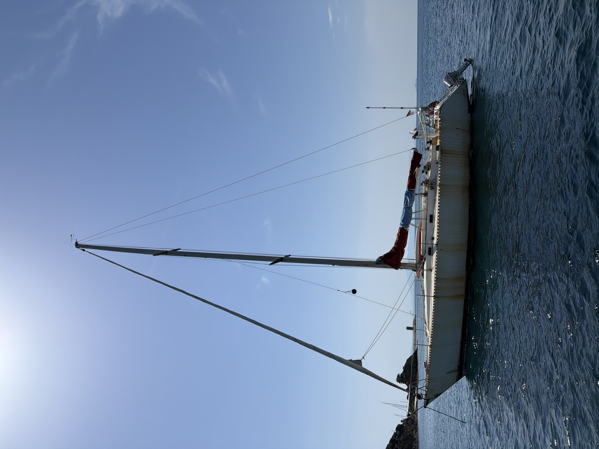 A white sailboat is anchored in the clear blue water off the coast of Punta Ala, Italy. The sailboat has a tall mast with sails furled, and an outboard motor is attached to the stern. The hull of the boat appears aged, with some rust stains visible. In the background, a rocky coastline with some vegetation is visible, along with a lighthouse. The sky is clear and bright with the sun overhead. The water is calm and a deep blue. There are no people visible on the boat or in the water.