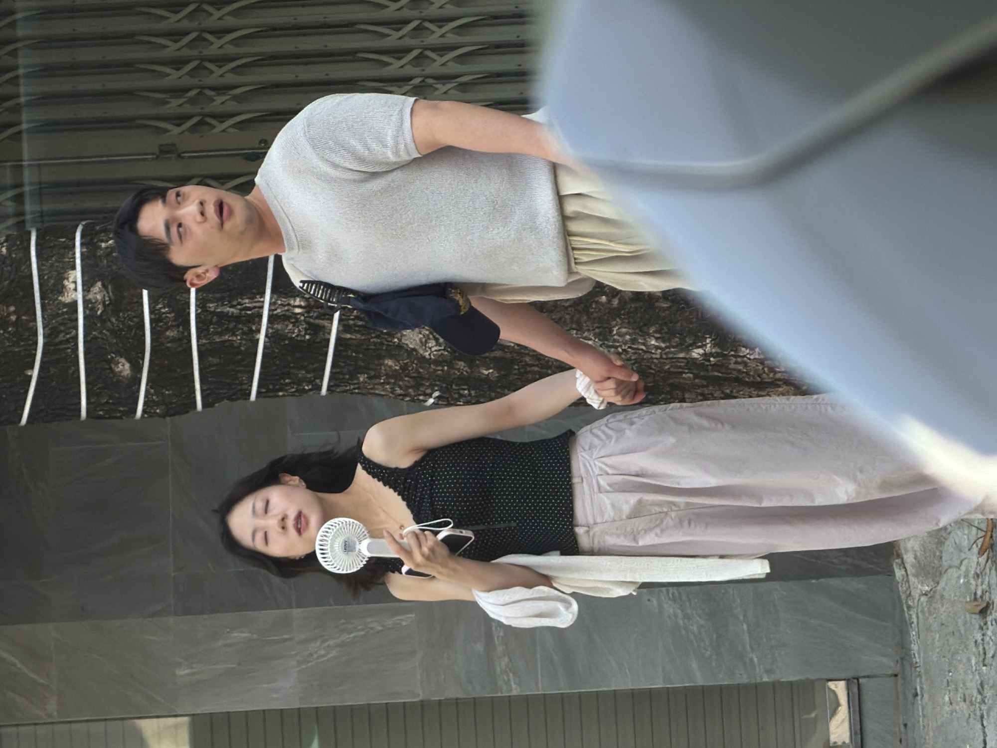 In Nha Trang Ward, Vietnam, a young woman with dark hair, wearing a black polka-dot tank top and light-colored wide-leg pants, holds a white portable fan in her right hand and a pink smartphone. A light-colored scarf is draped over her left arm. She is standing next to a man with short dark hair, wearing a light grey short-sleeved sweater and tan drawstring pants. He carries a dark bag slung over his left shoulder. They are holding hands, with the woman's left hand and the man's right hand clasped. They are positioned outdoors in front of a building with a dark, textured facade on the left and a tree trunk with white horizontal lines on the right. A metal gate is visible behind the man. The ground is paved and shows some debris. The foreground is partially obscured by a blurred light blue object.