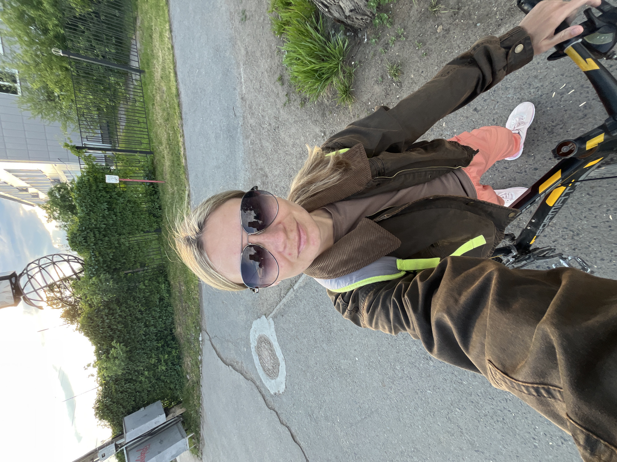 The image is a selfie taken by a woman riding a bicycle. She is wearing sunglasses, a brown jacket with a corduroy collar, a brown shirt, and salmon-colored pants. Her white sneakers are visible. The bicycle appears to be black with yellow accents. She is holding the handlebars.
The location appears to be a paved area, likely a sidewalk or bike path, with greenery, a fence, and buildings in the background. In the background left there is a large metallic sphere that can be found in Chaikovsky, Russia. There is a manhole cover on the ground near the woman.