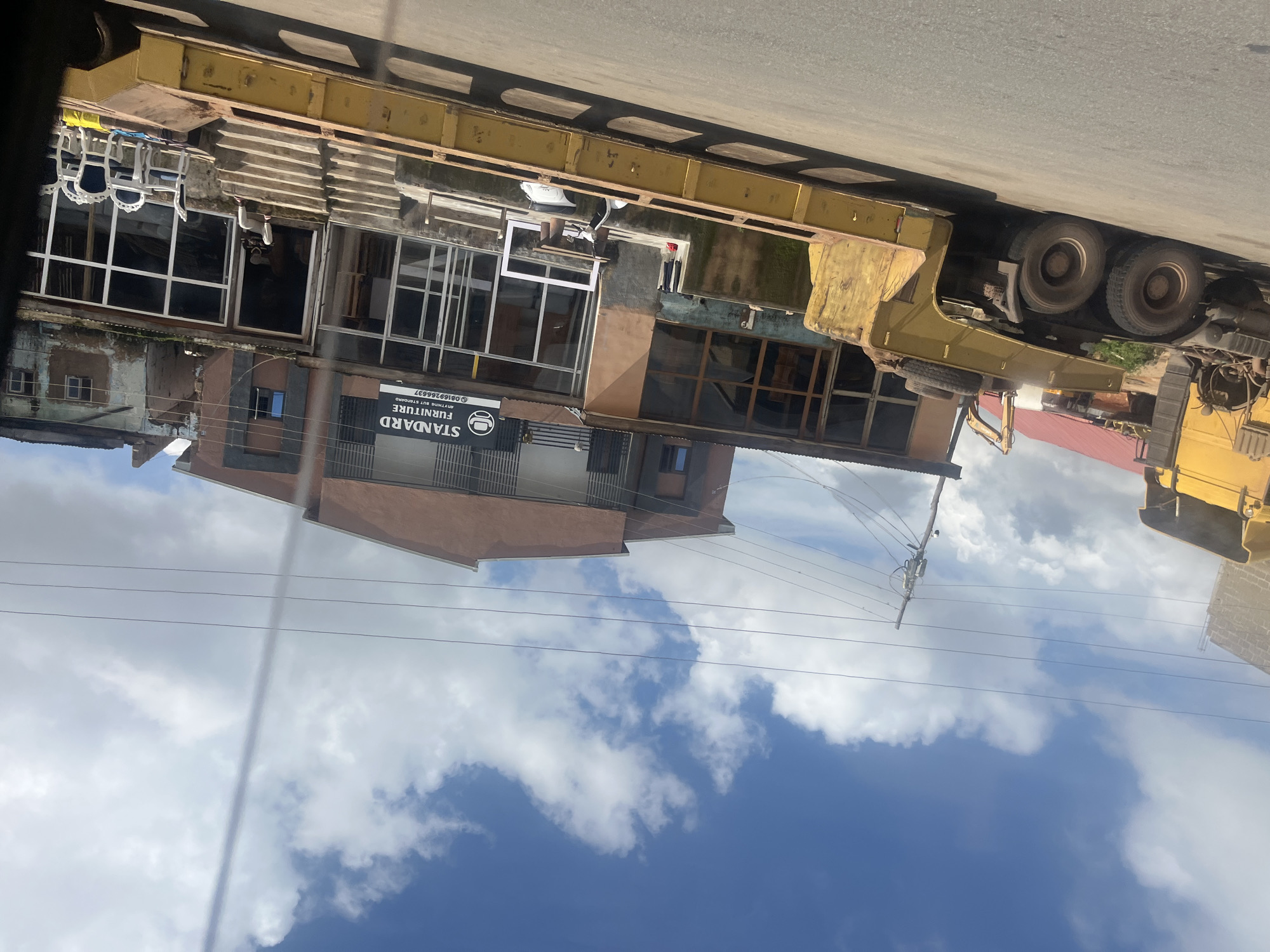 This is a ground-level, slightly angled, medium-shot photograph taken on a street in Jos, Nigeria, under a partly cloudy sky. In the foreground, a large, yellow, flatbed truck with multiple axles and large tires is parked on the asphalt road. The truck has a weathered appearance. Behind the truck, a two-story building with a brown facade and large glass windows is visible. A sign above the ground floor windows reads "STANDARD FURNITURE," with a phone number below it. The ground floor appears to be a storefront with furniture items displayed outside, including white chairs and a table. Steps lead up to the entrance of the building. A person is seated on the steps. To the left of the building, a portion of a yellow excavator is visible, and further back, a red-tiled roof can be seen. Power lines are strung across the scene. The overall impression is a commercial area with a heavy vehicle parked outside a furniture shop.