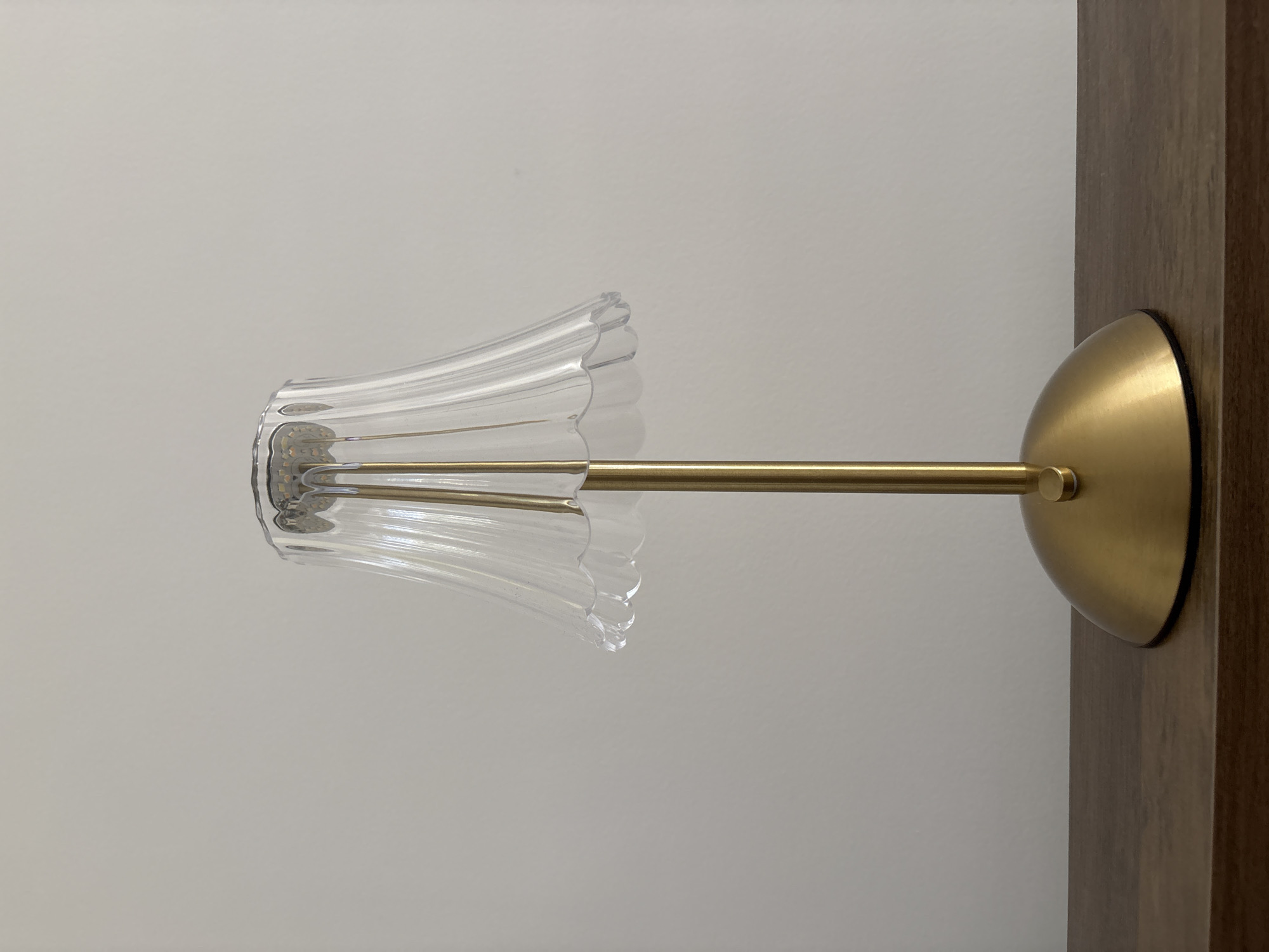 A gold desk lamp with a clear, fluted lampshade sits on a wooden surface against a plain, light-colored wall. The lamp has a circular base and a slender, vertical stem. The lampshade is wide at the top and tapers downwards, with a scalloped edge. The lamp is not illuminated. The image does not provide any information about its location in Liberec, Czechia.