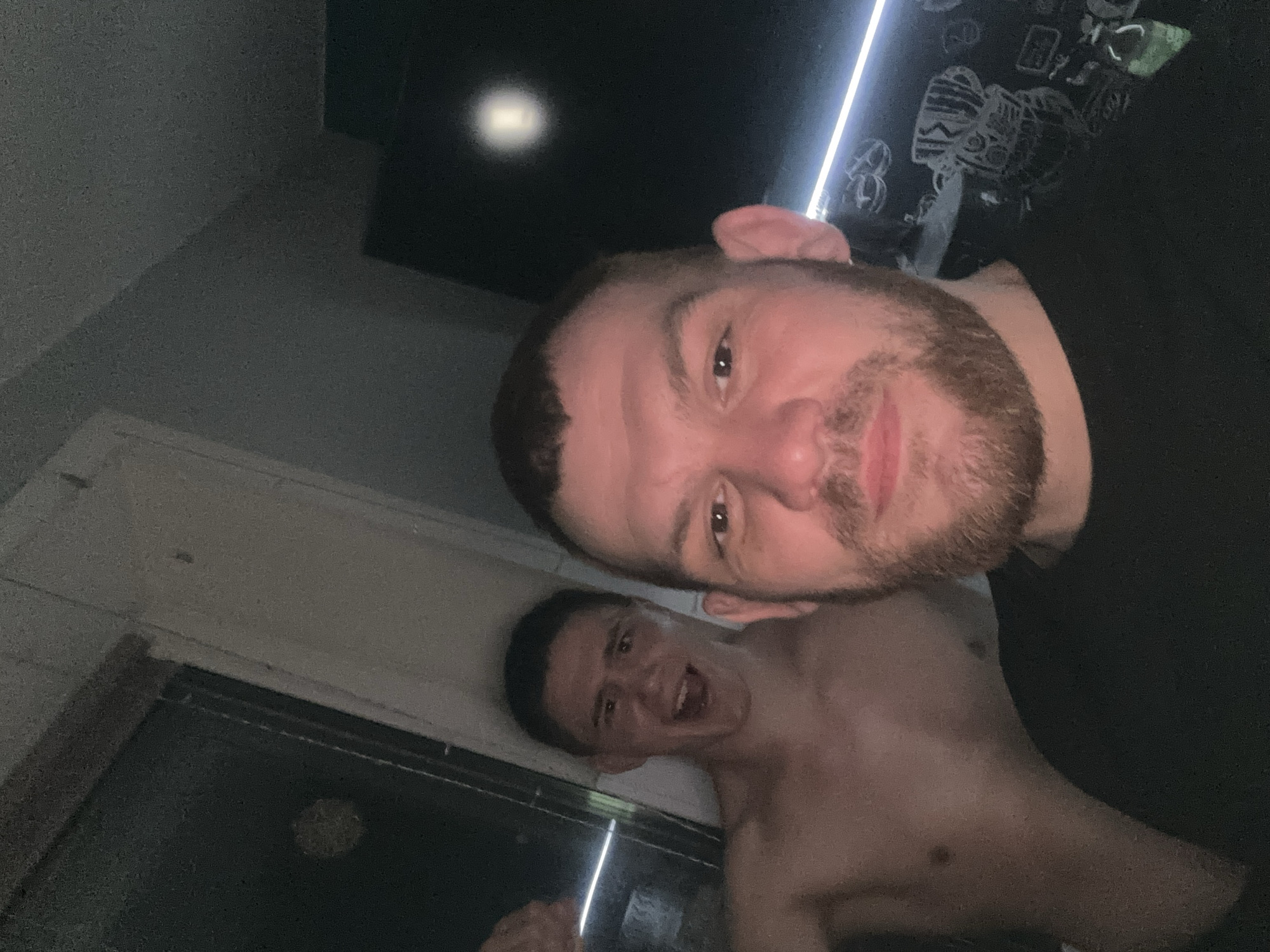 Two Caucasian males are shown in a dimly lit indoor environment. The older male, with a beard, is positioned closer to the camera, while the younger male, shirtless and appearing to be excited, is further in the background. The location appears to be in Perm, Russia. The background includes a window and a dark ceiling with decorative elements.