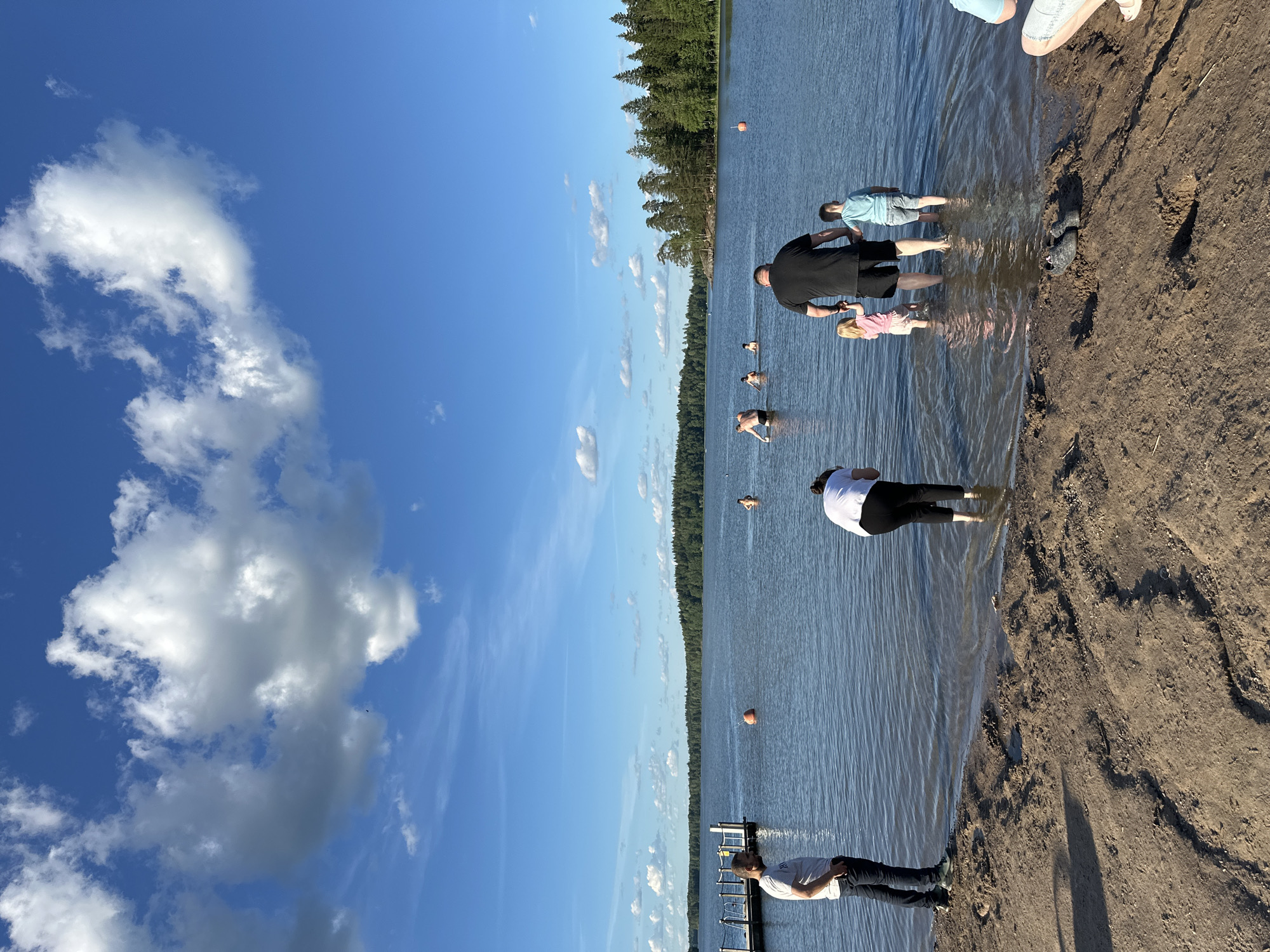 The image depicts a sunny day scene at a lake in the city of Unknown, Unknown. In the foreground, the sandy shore is visible, with a person's legs and feet partially submerged in the shallow water. Further into the lake, several people are swimming and wading. A man is standing on the shore, facing the water, while a woman in the foreground is bent over. In the mid-ground, a man and two children are standing in the water, with the man holding hands with both children. In the background, a dense forest lines the opposite shore of the lake. The sky is clear blue with scattered white clouds, and a wooden dock is visible on the left side of the image.