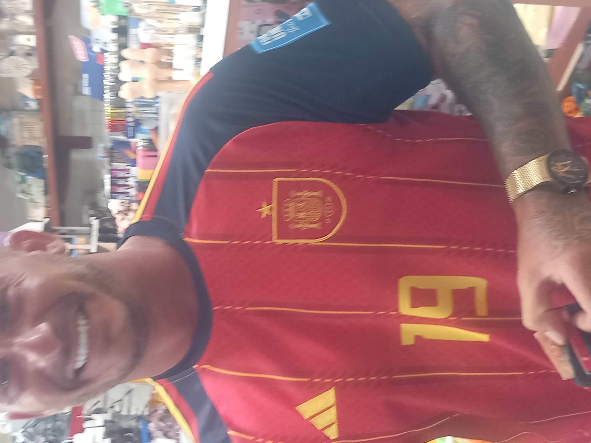 The image shows a man wearing a red and yellow striped Spain national football team jersey. The jersey has the Adidas logo on the left chest and the Spanish crest on the right chest. The number "19" is visible on the front of the jersey. The man has a gold watch on his left wrist and tattoos on his right arm. He is smiling. The background appears to be a retail store, with shelves stocked with various items. The exact location within the city of Kos, Greece, cannot be determined from the image alone.