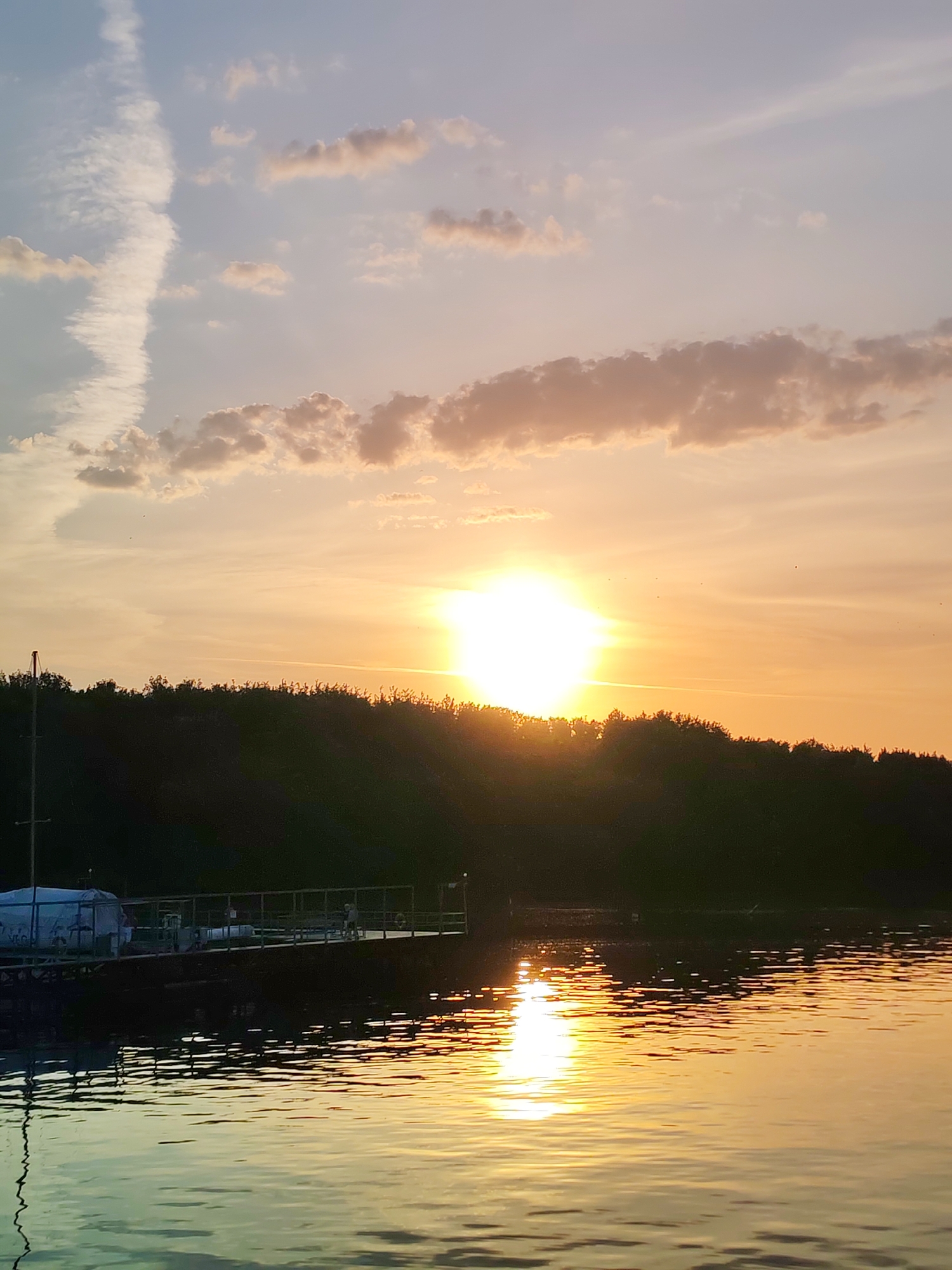 The image depicts a sunset over a body of water. In the foreground, there is a pier or dock with a structure on it. The structure appears to be covered or enclosed. A dark, silhouetted treeline stretches across the middle of the frame, with the sun setting behind it. The sun is bright and creates a reflection on the water's surface. The sky is a mix of orange, yellow, and blue hues with scattered clouds. The water shows ripples and reflections of the sky and the sun's light. The location is most likely Ermolino, Russia.