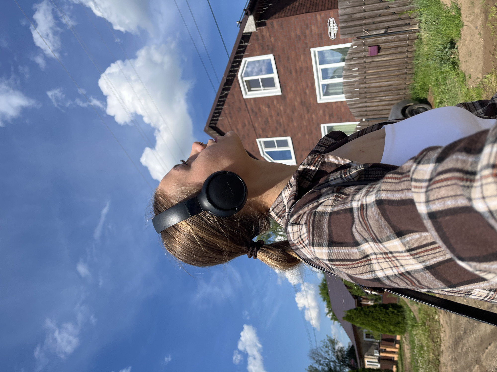 A young woman is captured in a selfie taken outdoors, looking upwards towards a bright blue sky with scattered white clouds. She is wearing black over-ear headphones and a brown and white plaid shirt over a white top. Her hair is tied back in a ponytail.
In the background, a two-story brick house with white-framed windows is visible, along with a wooden fence and green grass. The scene suggests a sunny day, likely in the afternoon, given the lighting. The overall mood appears relaxed and contemplative, as the woman seems to be enjoying the outdoors and her music. The image is oriented vertically. No visible text is present in the image.