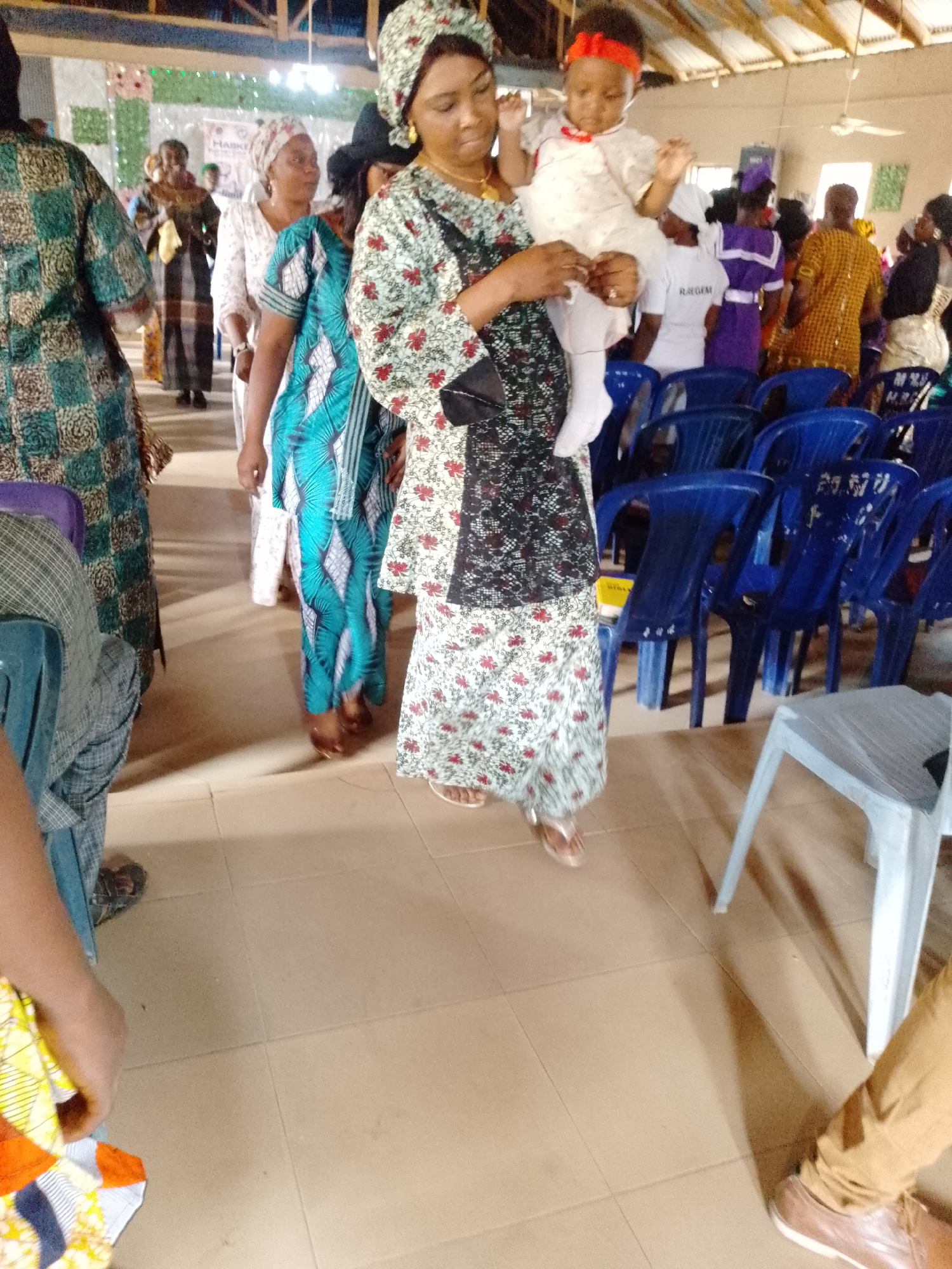 The image shows an indoor gathering, likely in a church or community hall, based on the presence of rows of blue plastic chairs and the attire of the individuals. A woman, wearing a patterned dress and matching headwrap, is holding a baby in a white dress with a red headband. Other people are walking in the background, some in patterned clothing and head coverings. The floor is tiled. The location appears to be a building in an unspecified city.