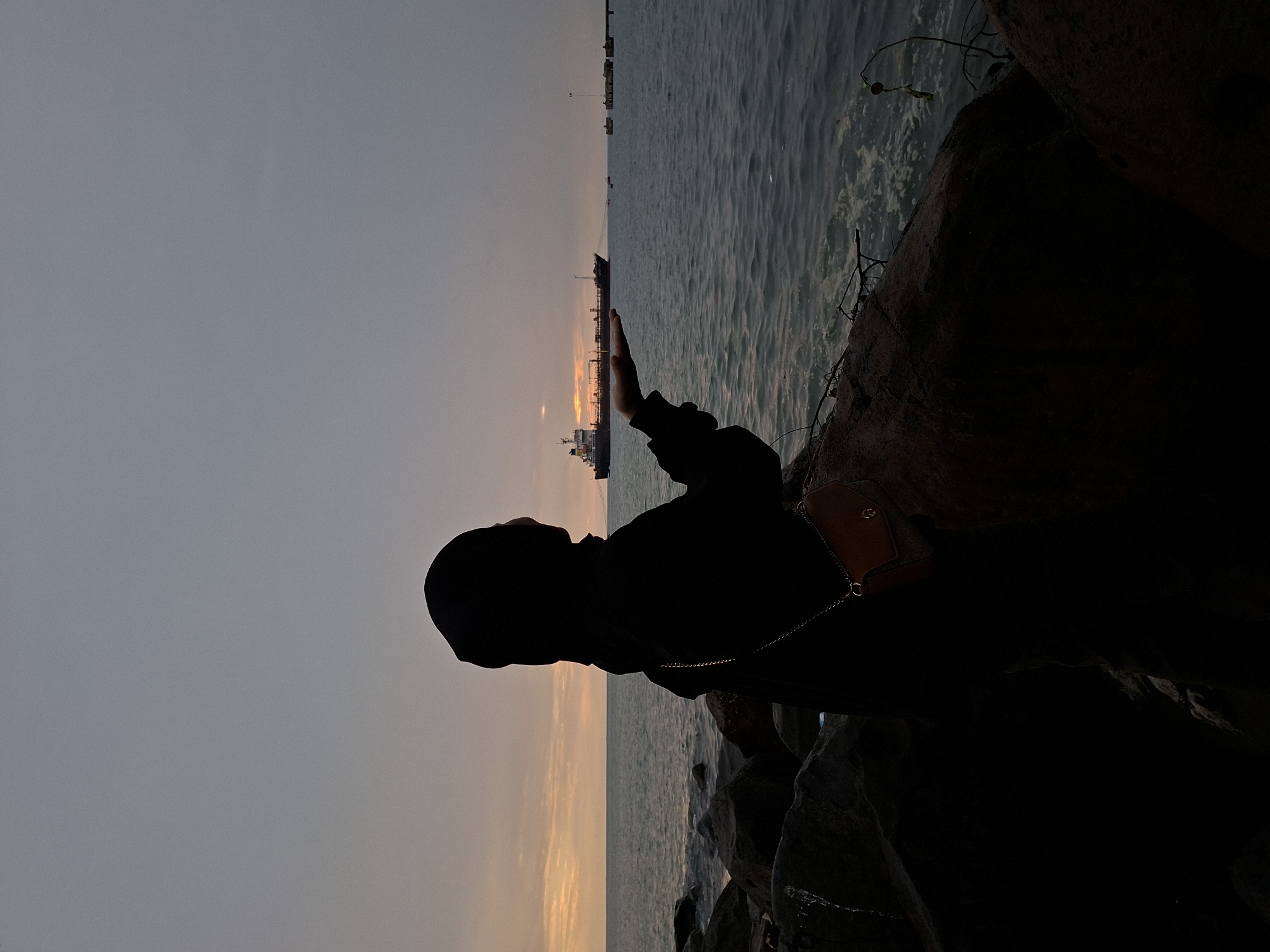 A silhouetted woman wearing a hijab stands on a rocky shore facing the ocean at dusk. Her right hand is extended forward, palm up, as if to hold something. In the distance, a large cargo ship sails on the water. The sky is overcast with soft hues of orange and gray from the setting sun. The foreground is dominated by dark, wet rocks, with the sea lapping at their edges. The scene appears to be at Ujung Blang Beach, Lhokseumawe, Indonesia, a popular spot for sunset views.