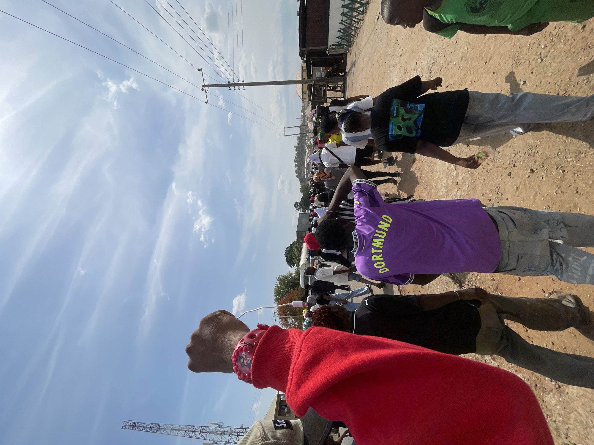 A large group of people is walking along a dirt road in an outdoor environment. An individual in the foreground raises their fist. The weather appears sunny, with a blue sky containing scattered clouds. The location may be the city of Lokoja, Nigeria. Power lines and poles are visible above the heads of the crowd. A red sweatshirt is visible in the foreground. A purple shirt with the word "DORTMUND" printed on the back can be seen. Other clothing includes a green shirt, white shirts, and black shirts. A construction crane can be seen in the background.