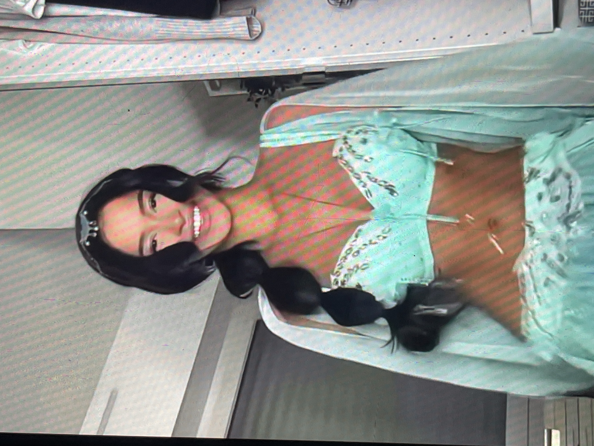 A young woman with dark hair styled in a long braid is wearing a turquoise, ornate costume, resembling that of Princess Jasmine from Aladdin. She has a jeweled headband and a necklace. She is standing in what appears to be an indoor environment, possibly a closet or a dressing room, with clothing hanging in the background on the right and a door on the left. The lighting is somewhat harsh, with visible scan lines across the image, suggesting it may be a photograph of a screen. The woman is smiling broadly.