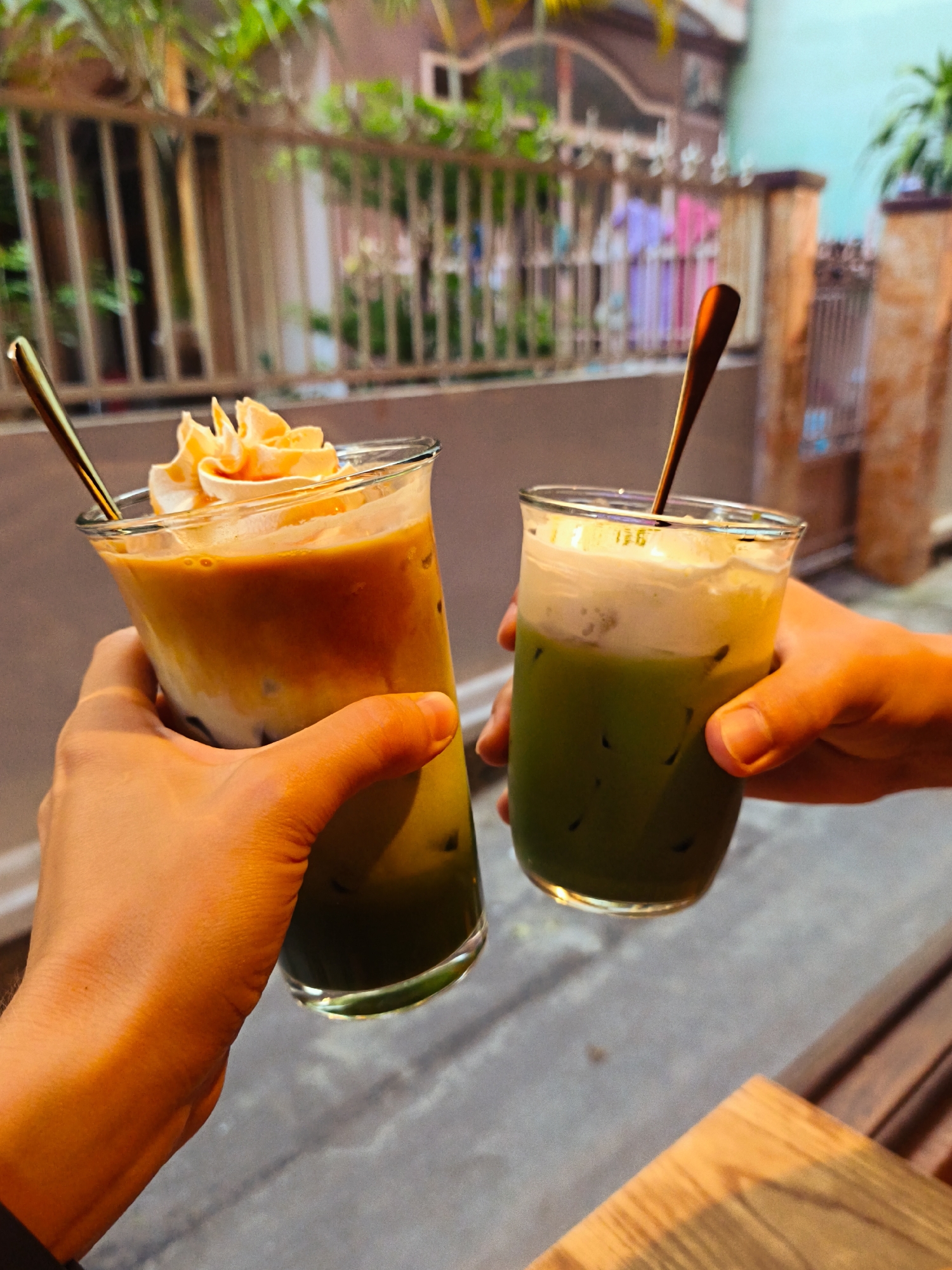 Two hands hold clear glasses of iced beverages. The glass on the left contains a layered drink with a light brown top, possibly coffee with cream, topped with whipped cream. A gold spoon rests inside. The glass on the right holds a green beverage, likely iced matcha, with a layer of white foam on top, also with a gold spoon. Both drinks have ice cubes. The scene appears to be outdoors, with a fence and foliage in the background, suggesting a street or patio setting in the city of Bac Nha Trang Ward, Vietnam. The lighting indicates it may be late afternoon or early evening.