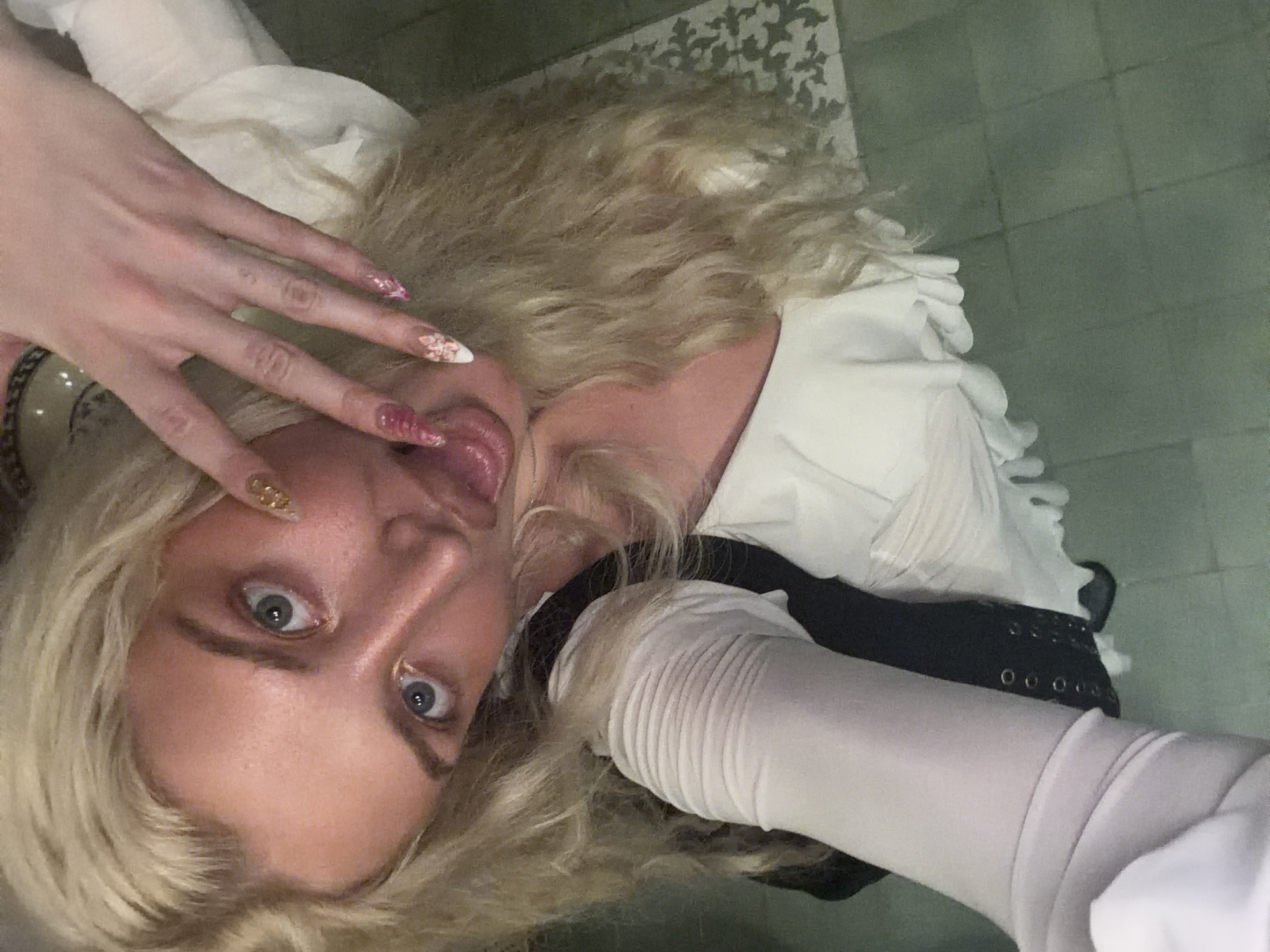 A close-up, rotated portrait features a person with blonde, wavy hair and pale skin, looking directly at the camera with wide, expressive blue-grey eyes. The image is rotated approximately 90 degrees counter-clockwise, positioning the subject's head towards the upper right.
The person's lips are slightly open, with their tongue playfully extended. One hand is raised near their face, showcasing long, elaborate artificial nails adorned with pink glitter, white tips featuring floral designs, and golden accents. The other arm, visible on the left side of the frame, is covered by a fitted white glove or sleeve with a ribbed texture, and appears to be bent near their head.
The subject is wearing a white garment with visible layers or ruffles, suggesting a dress or top. Beneath this, a black garment with metallic eyelets, possibly a corset or belt, can be seen. The background features a dark green or grey tiled surface, likely a floor, and a darker patterned area, suggesting an indoor setting illuminated by artificial light. The overall expression is playful and engaging. No visible text is present. While the location context provided is North Kuta, Indonesia, the image itself does not contain specific visual cues to confirm this, beyond being an indoor scene.