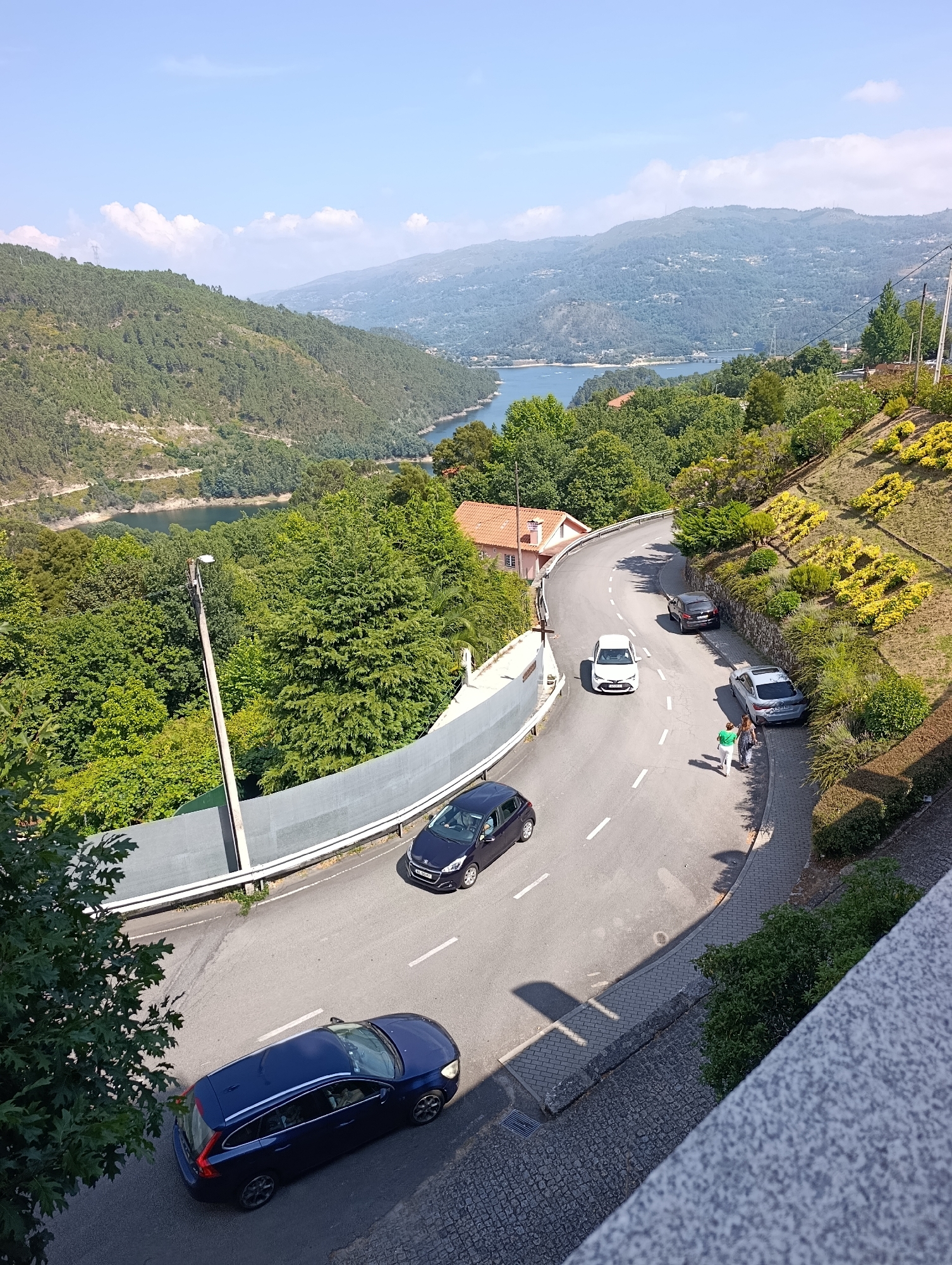 This aerial, wide-angle view captures a scenic, winding road in Terras de Bouro, Portugal, flanked by lush green trees and steep, terraced hillsides. The road curves around a hillside, with several cars parked and driving on it. In the foreground, a dark blue station wagon is parked on the lower part of the road. Further up, a dark colored SUV is parked, and a silver sedan is parked next to it. Two people, a woman in a green shirt and a woman in a black and white dress, are walking on the sidewalk next to the silver sedan. A white sedan is driving uphill. In the background, a wide river or lake is visible, with rolling, tree-covered hills in the distance. The sky is clear and blue with scattered white clouds. The environment appears to be rural and natural, with a focus on the road and its surroundings.