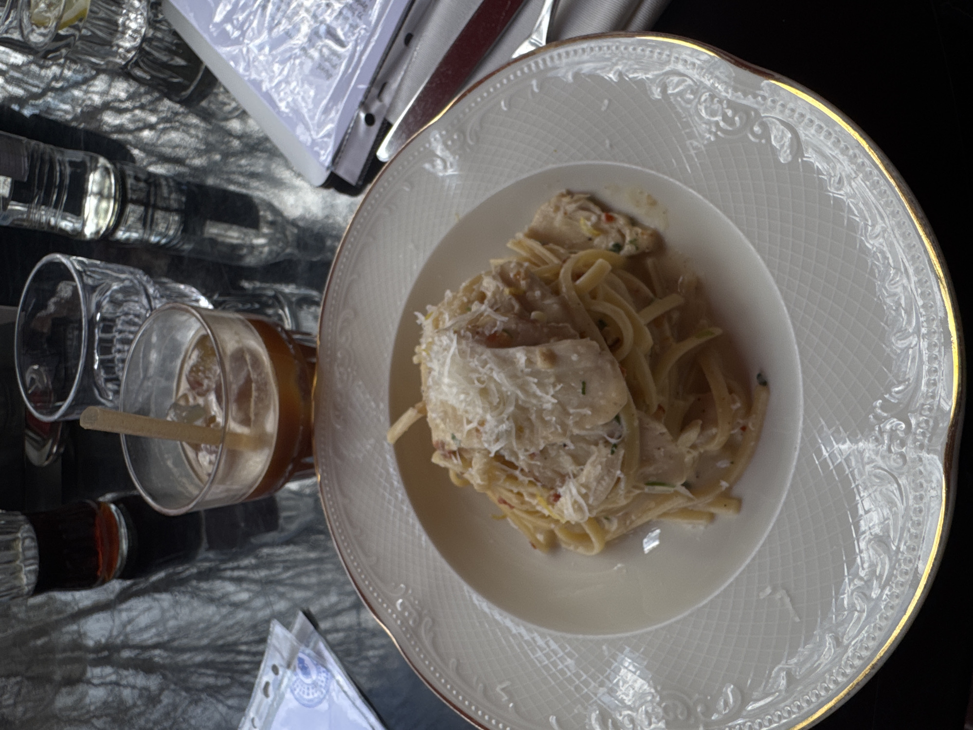 A high-angle shot captures a dining table at Louis Charden, Yerevan, Armenia. The central item is a white, ornate dinner plate featuring a gold rim and a detailed embossed pattern. It holds a portion of linguine pasta coated in a creamy sauce, mixed with chicken pieces, and generously topped with grated cheese. Surrounding the plate on the dark, reflective table surface are several items: a clear glass containing an iced dark brown beverage with a light brown straw, an empty textured clear glass, a clear plastic water bottle, and a clear glass of water with a visible lemon slice. A smaller, dark glass with a dark liquid is also present. The table's reflective surface prominently displays silhouettes of tree branches, suggesting an outdoor dining area or an indoor space with large windows overlooking foliage. To the left, a white document with a blue circular logo is partially visible, protected by a clear plastic sleeve. To the right, a white menu or document is visible within a clear plastic sleeve, alongside a metal knife with a dark handle resting on a folded white napkin. No individuals are present in the frame.