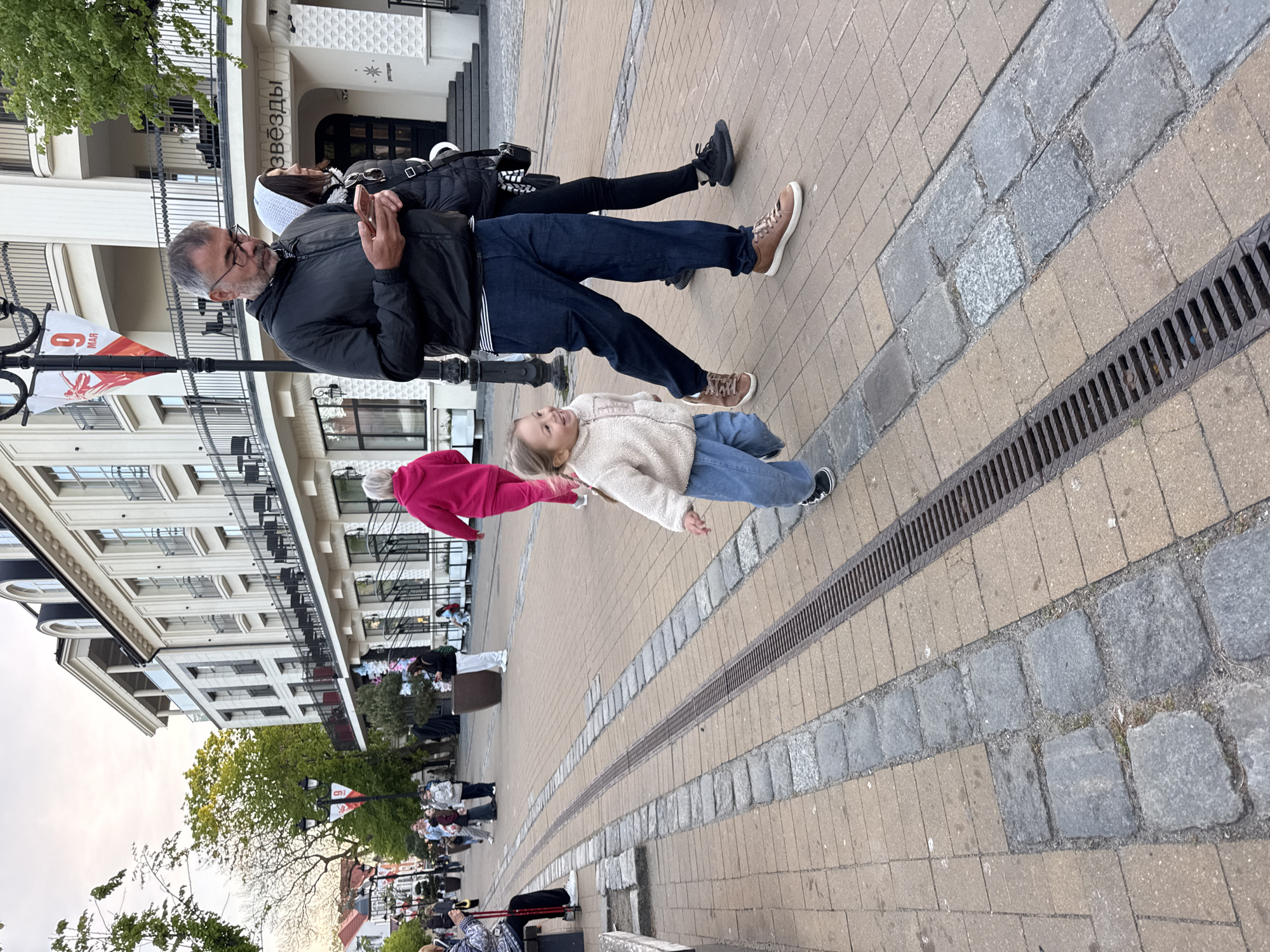 A family is walking on a paved sidewalk near a building in Zelenogradsk, Russia. An adult male is using a smartphone; a second adult and a young child walk alongside him. The pavement is composed of rectangular brick tiles interspersed with rows of grey cobblestones and narrow drainage grates. A building with white walls and black iron balconies is visible in the background, bearing the name "Звезды" (Stars). A flag with the date "9 MAR" is attached to a lamp post. The sky is overcast, and other pedestrians are visible in the distance.