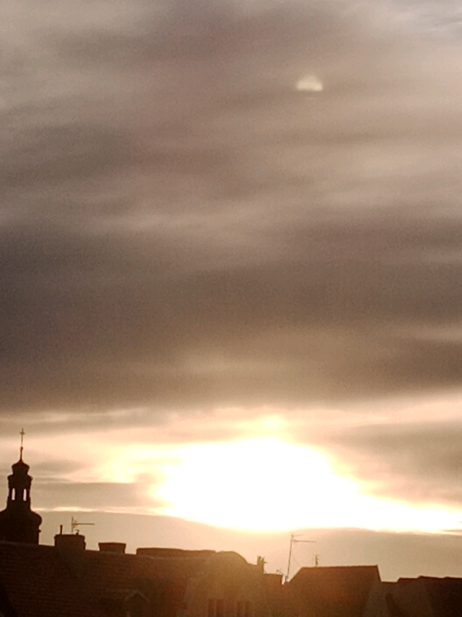 The image depicts a sunset over a European city, possibly Bydgoszcz, Poland. The sky is filled with hazy, muted tones of orange, pink, and grey, with the sun obscured by clouds, creating a diffused glow. In the foreground, silhouetted against the bright sky, are the rooftops of buildings. A prominent church steeple with a cross on top is visible on the left side of the frame. The architecture suggests an older part of a city, with a mixture of rooflines and chimney structures. The overall impression is one of a calm, atmospheric evening.
