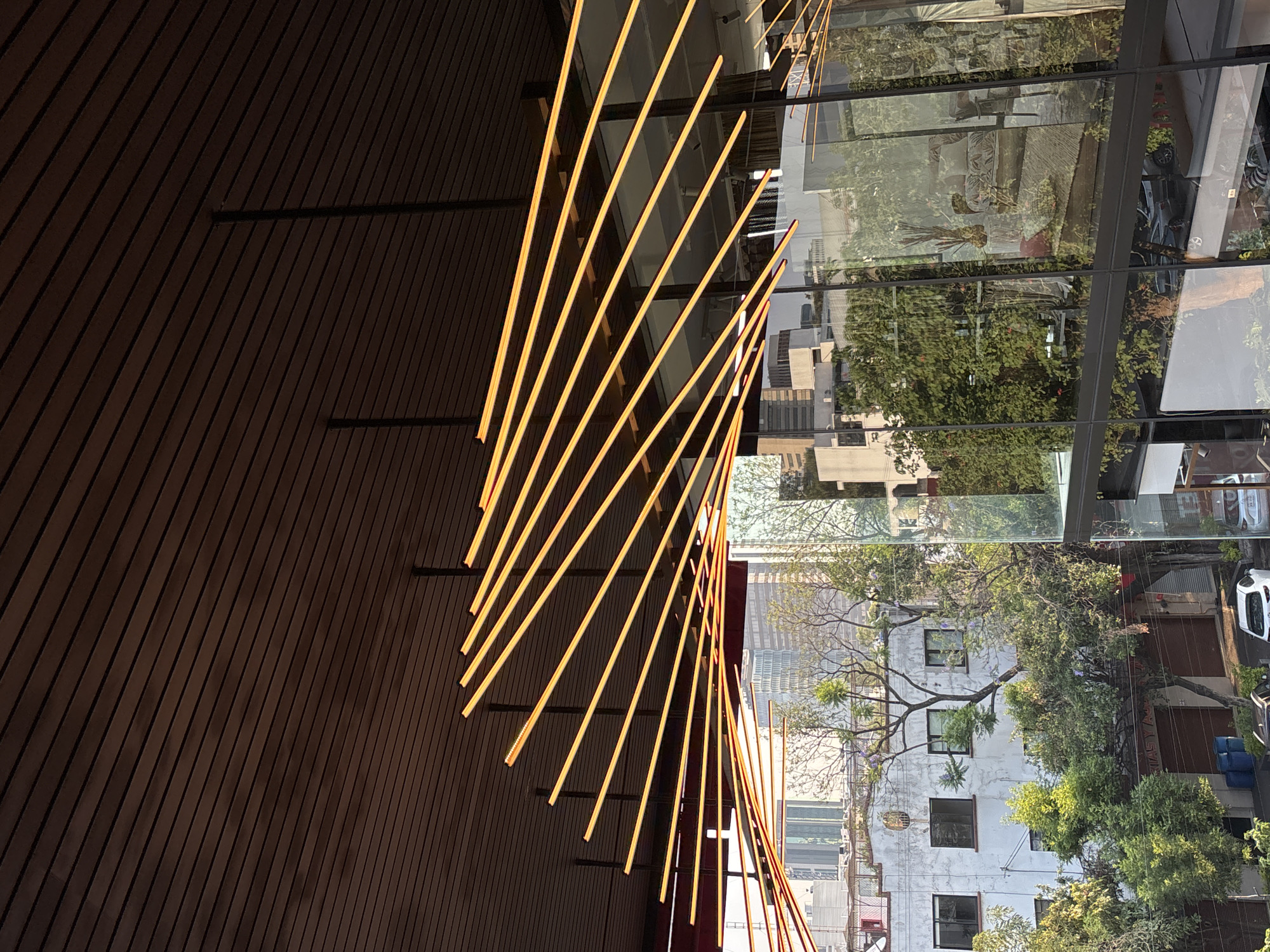 The image captures a modern architectural detail in Mexico City, Mexico. A facade of dark wood planks runs vertically along the left side of the frame, punctuated by a series of parallel, evenly spaced, horizontally oriented black metal beams. Attached to these beams are evenly spaced illuminated golden rods that radiate out from the wood facade. To the right of the wood facade is a large expanse of glass panels that reflect an exterior cityscape. The reflections show buildings, trees, and signs, all contributing to the urban environment. The lighting suggests an overcast day, with diffuse sunlight illuminating the golden rods and reflecting off the glass. The composition is tightly cropped, emphasizing the geometric patterns of the architectural elements and the reflected urban scene.