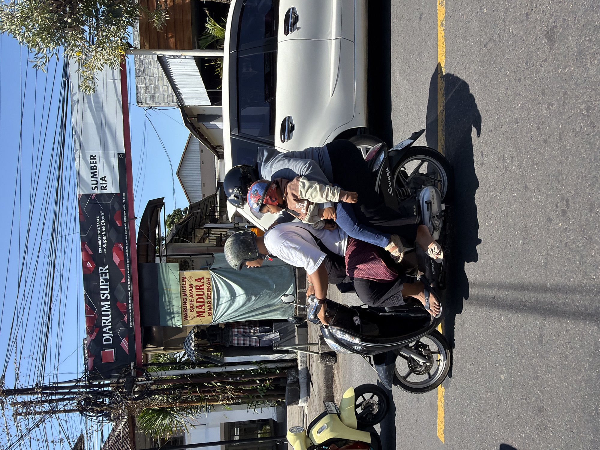 The image depicts a street scene in an urban environment under a bright, sunny sky. A black scooter carrying three individuals is the central focus, traveling on a paved road with a yellow line. The front rider, a man wearing a white shirt and a grey helmet, is driving. Behind him, a woman in a grey hoodie holds a young child wearing a red and blue helmet. The child is dressed in a light brown sweatshirt and blue pants. The scooter has "Scoopy" written on its side, and the make "HONDA" is visible on the engine. To the left of the scooter, a yellow scooter is parked. In the background, there is a white van, several buildings with tiled roofs, and a large banner overhead. The banner contains text in Indonesian, advertising "DJARUM SUPER" and "SUMBER RIA," as well as a warning about smoking. Another sign for "WARUNG MUSLIM SATE AYAM MADURA SINAR BERKAH" is also visible. Overhead, a complex network of electrical wires crisscrosses the scene. The overall atmosphere suggests a typical day in a bustling city.