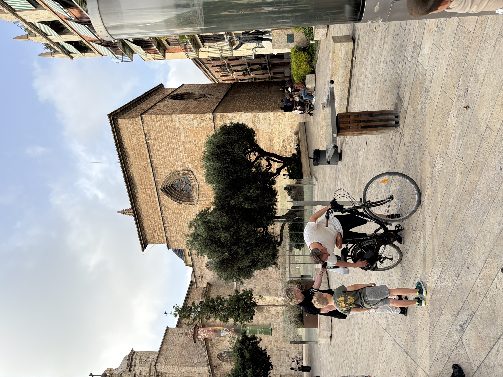 In an outdoor urban plaza, a man in a white t-shirt is leaning over a black bicycle, appearing to adjust it. A woman in a black t-shirt and a young boy in a green t-shirt and grey shorts stand beside him, looking at the bicycle. The plaza is paved with light-colored stone tiles. Behind the individuals, a large, old stone building with arched windows and a domed section is visible. To the right of this building, a modern multi-story building with balconies and striped awnings stands. Further to the right, a dark statue on a pedestal is visible. Trees, including a large, old olive tree, are planted in the plaza. Other people are seen in the background, some sitting on benches and some walking. The sky is cloudy.