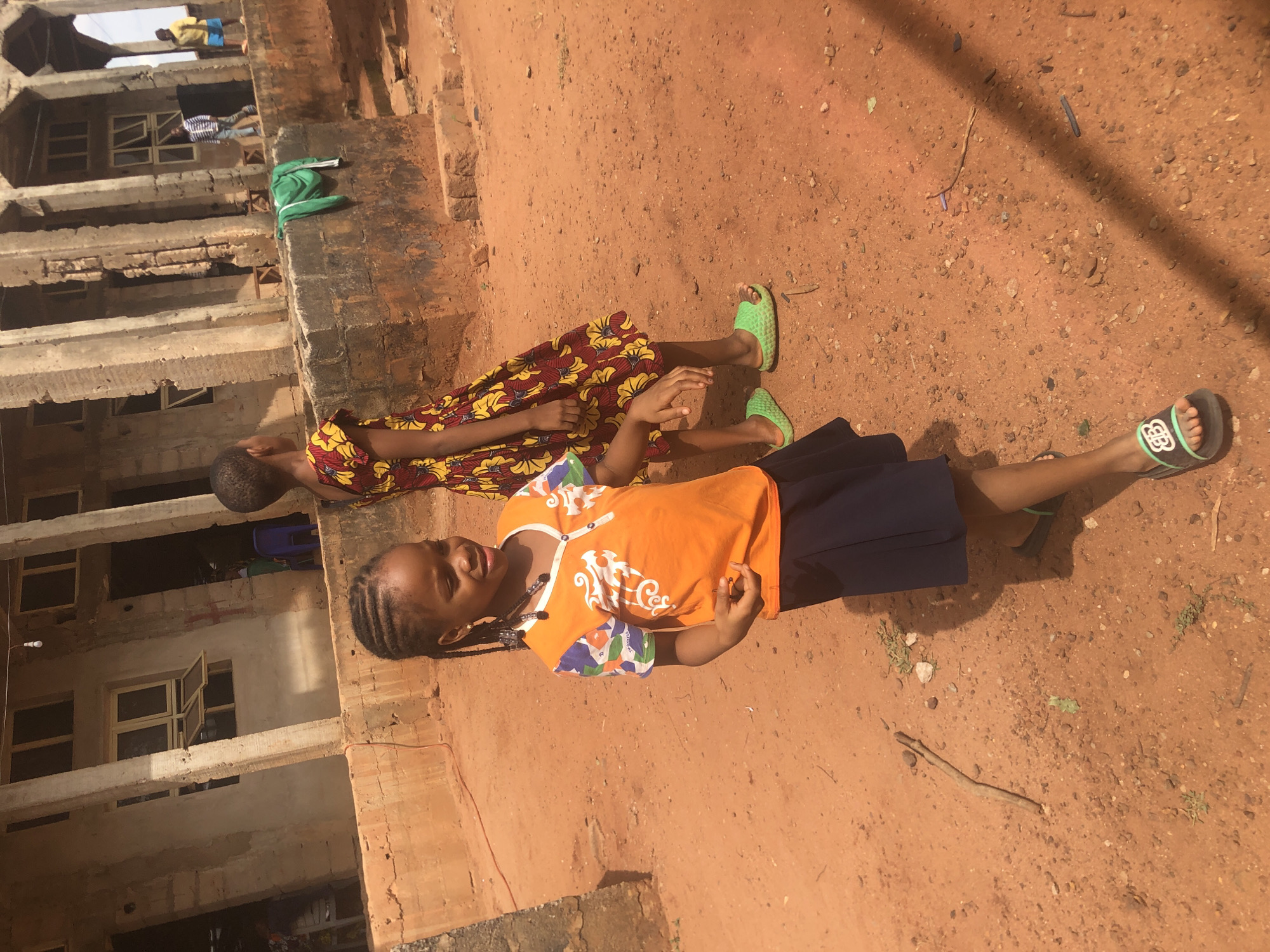Two young girls lie on red dirt in an outdoor setting in Nteje, Nigeria. The girl closest to the viewer is wearing an orange top with blue and white patterned sleeves, a navy skirt, and green and black sandals. She is lying on her back and looking to the right of the frame. The second girl is wearing a dress with a yellow and red floral pattern and green sandals, lying on her side. A partially constructed building with exposed concrete and brick is behind and to the left of the girls. The ground is covered in red dirt with small rocks and debris.