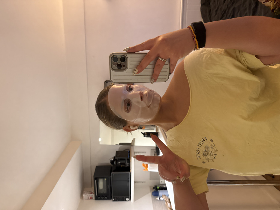 A person with a sheet mask on their face takes a selfie, holding their phone up with one hand and flashing a peace sign with the other. They wear a yellow t-shirt. A fitness tracker and bracelets adorn their left wrist. The background reveals a mirror reflecting the person's bathroom, including a microwave and small refrigerator on the counter. The lighting appears to be natural, casting a soft glow throughout the scene.