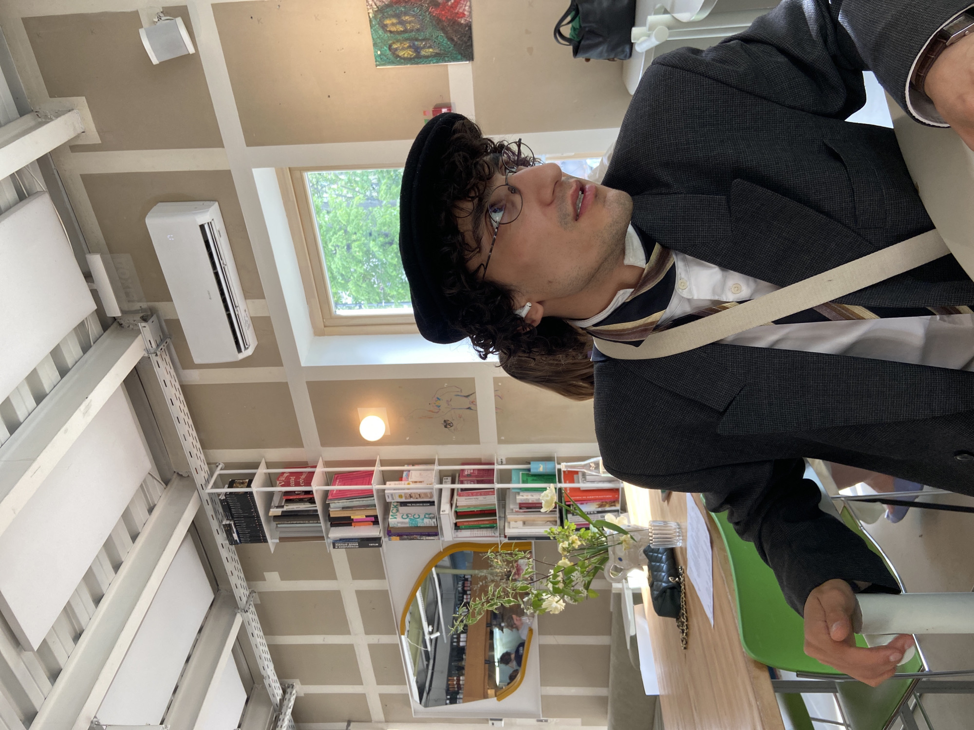 The image shows a person wearing a black beret, glasses, a white shirt, a striped tie, and a gray suit jacket. They appear to be seated at a table, possibly in a cafe or restaurant. In the background, there is a bookshelf filled with books, a mirror, and a vase with flowers. There's also a window with a view of greenery. The ceiling has a grid-like structure, with what looks like acoustic panels and an air conditioning unit. I am unable to identify the specific location in Moscow.