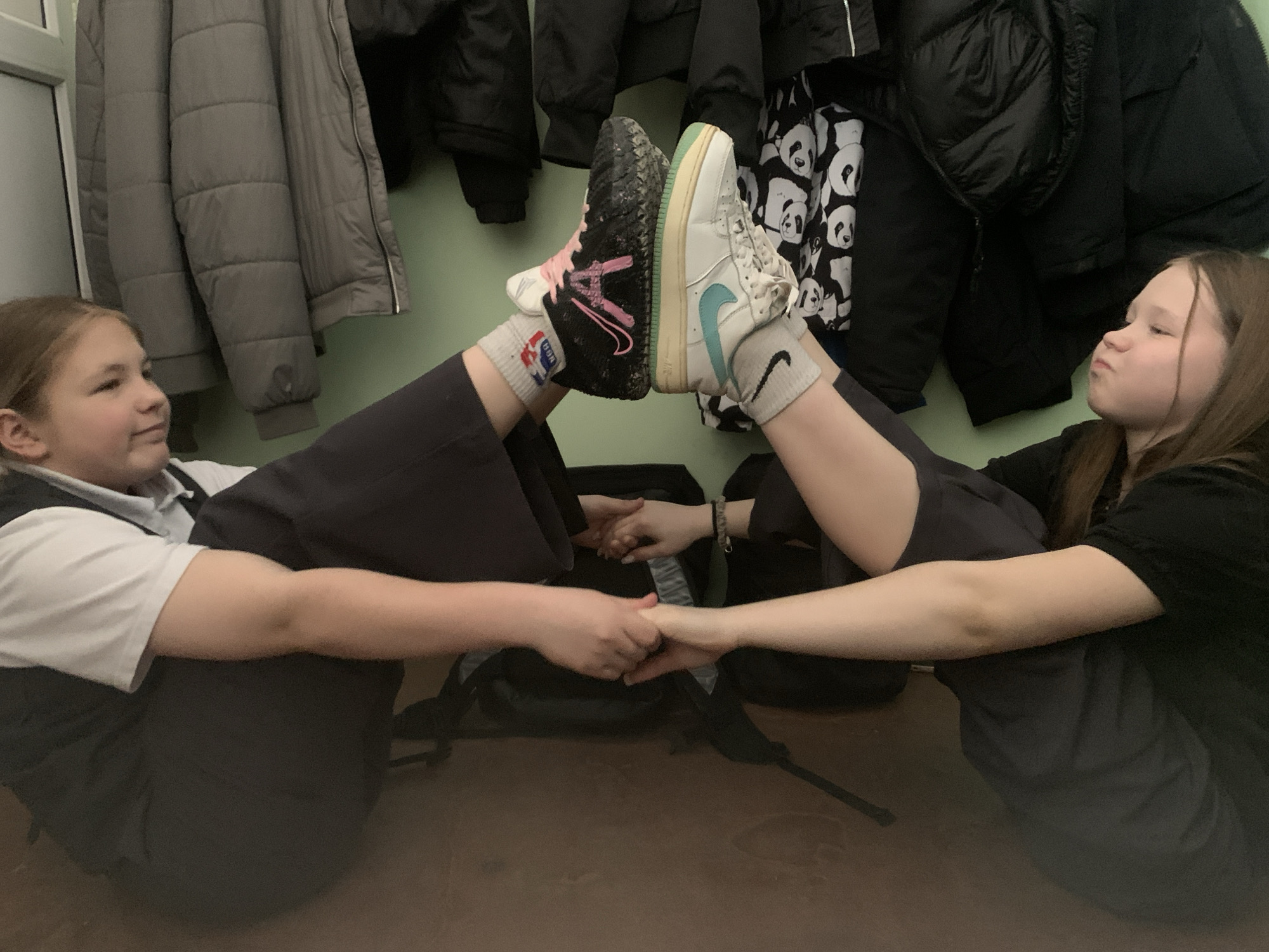 Two girls are sitting on the floor facing each other, with their legs raised and feet touching. They are holding hands. The girl on the left has brown hair and is wearing a white shirt with a black vest. The girl on the right has brown hair and is wearing a black shirt. Both girls are wearing dark pants. The girl on the left is wearing black and white sneakers with pink laces, and the girl on the right is wearing white sneakers with a green swoosh. Behind them is a wall with several coats and jackets hanging on it. In between them are backpacks.
Based on information provided about the location and content analysis, it is likely that this image was taken in the city of Chaikovsky, Russia.