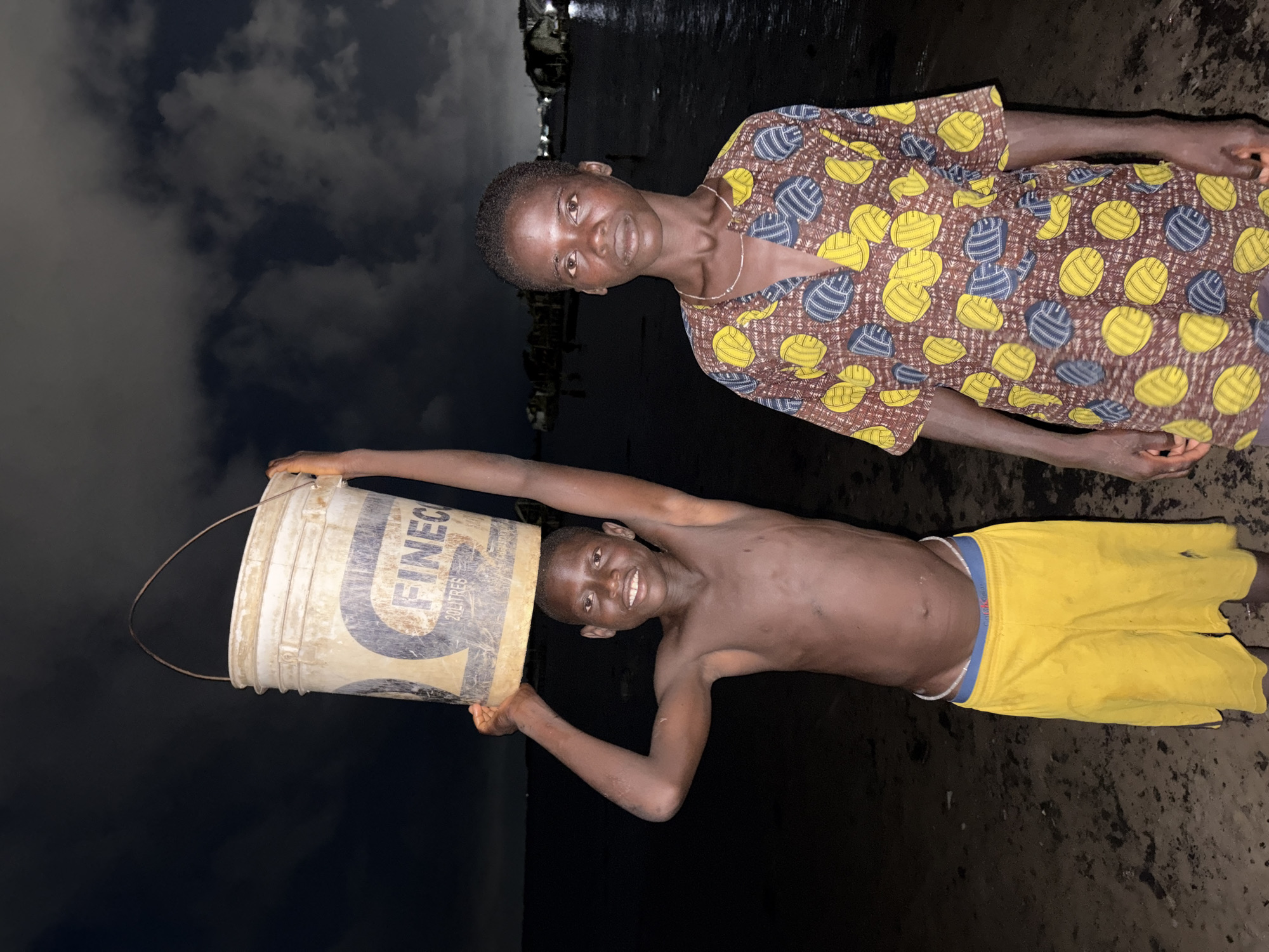 Two individuals are shown in an outdoor setting, likely a beach, possibly near Ikoyi, Nigeria, based on the subjects' appearances. The foreground features a shirtless youth in yellow shorts holding a bucket over his head. The bucket has the word "Finec" printed on its side. Behind him stands another individual wearing a patterned top. The background is dark and suggests a nighttime scene with a body of water and some structures visible in the distance. The sky is cloudy.