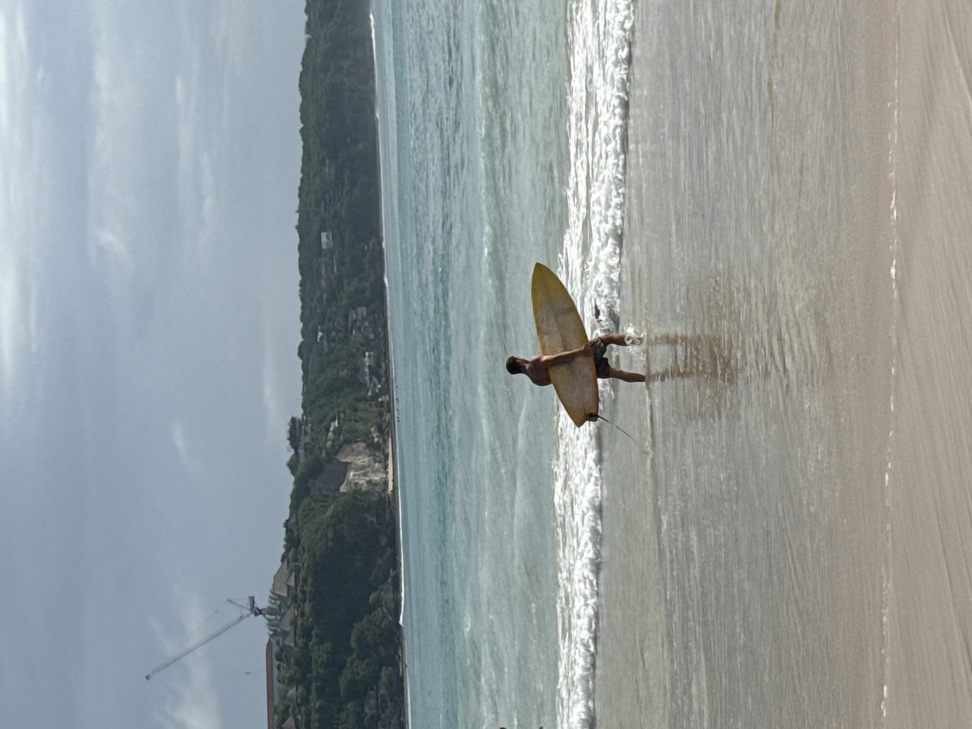 A man stands in the shallow surf of a beach in Kuta Selatan, Indonesia, holding a surfboard. The beach stretches to the right, with a line of waves breaking along the shore. The water is a pale turquoise. The shoreline on the left is covered with dense green foliage and buildings. A tall construction crane is visible in the distance on the left. The sky is overcast.