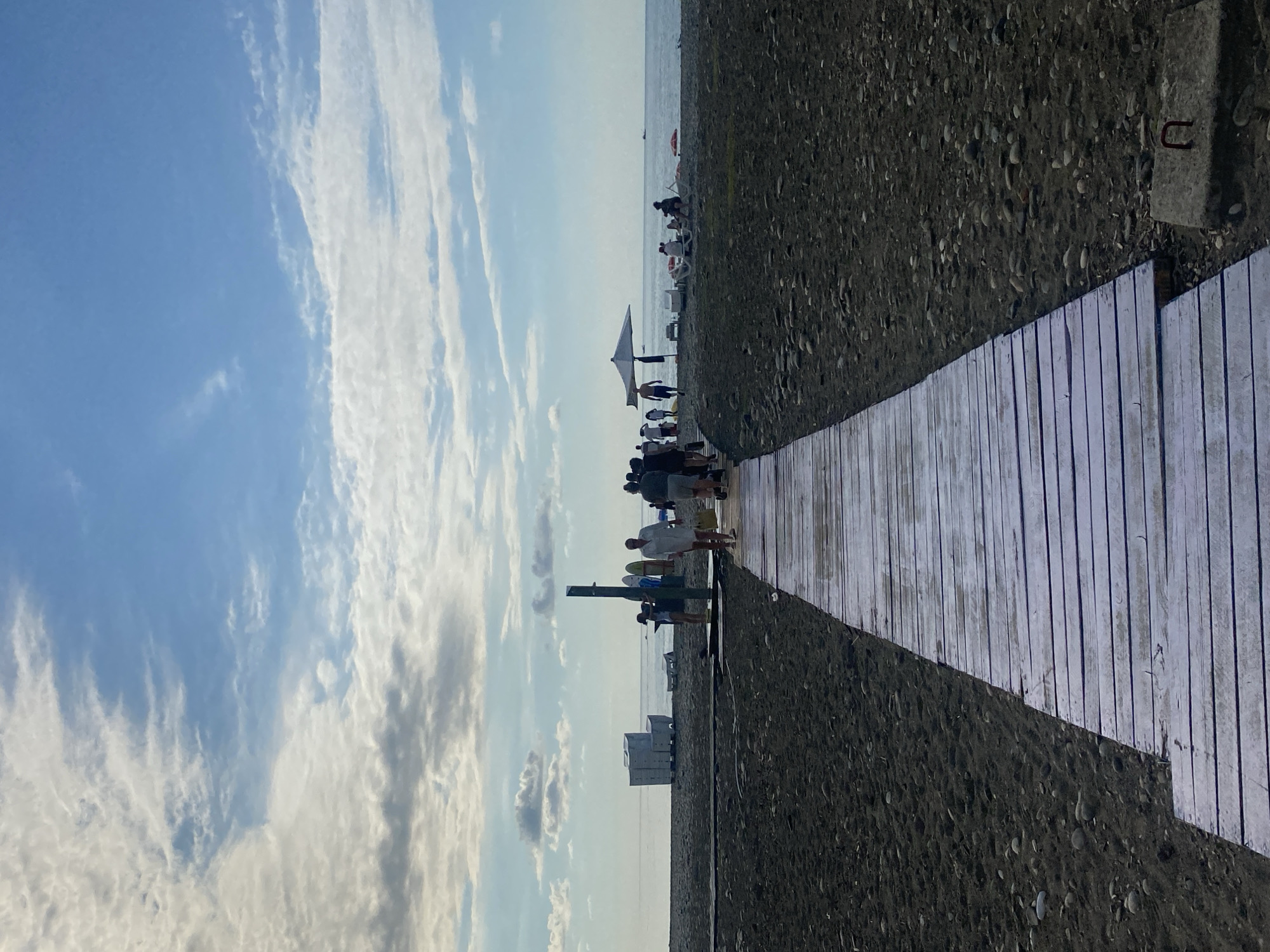 The image depicts a wooden boardwalk leading towards the Black Sea at a beach in Batumi, Georgia. The boardwalk is made of weathered, light-colored planks and is wet, suggesting recent rain or spray from the sea. The beach itself is composed of dark sand and pebbles. In the distance, people are visible walking along the beach and near the water's edge. Some are seated under beach umbrellas. There are lounge chairs, surfboards, and what appear to be stacked white chairs on the sand. The sea is calm, meeting the horizon under a partially cloudy sky with hues of blue and white. The scene captures a typical day at a seaside resort.